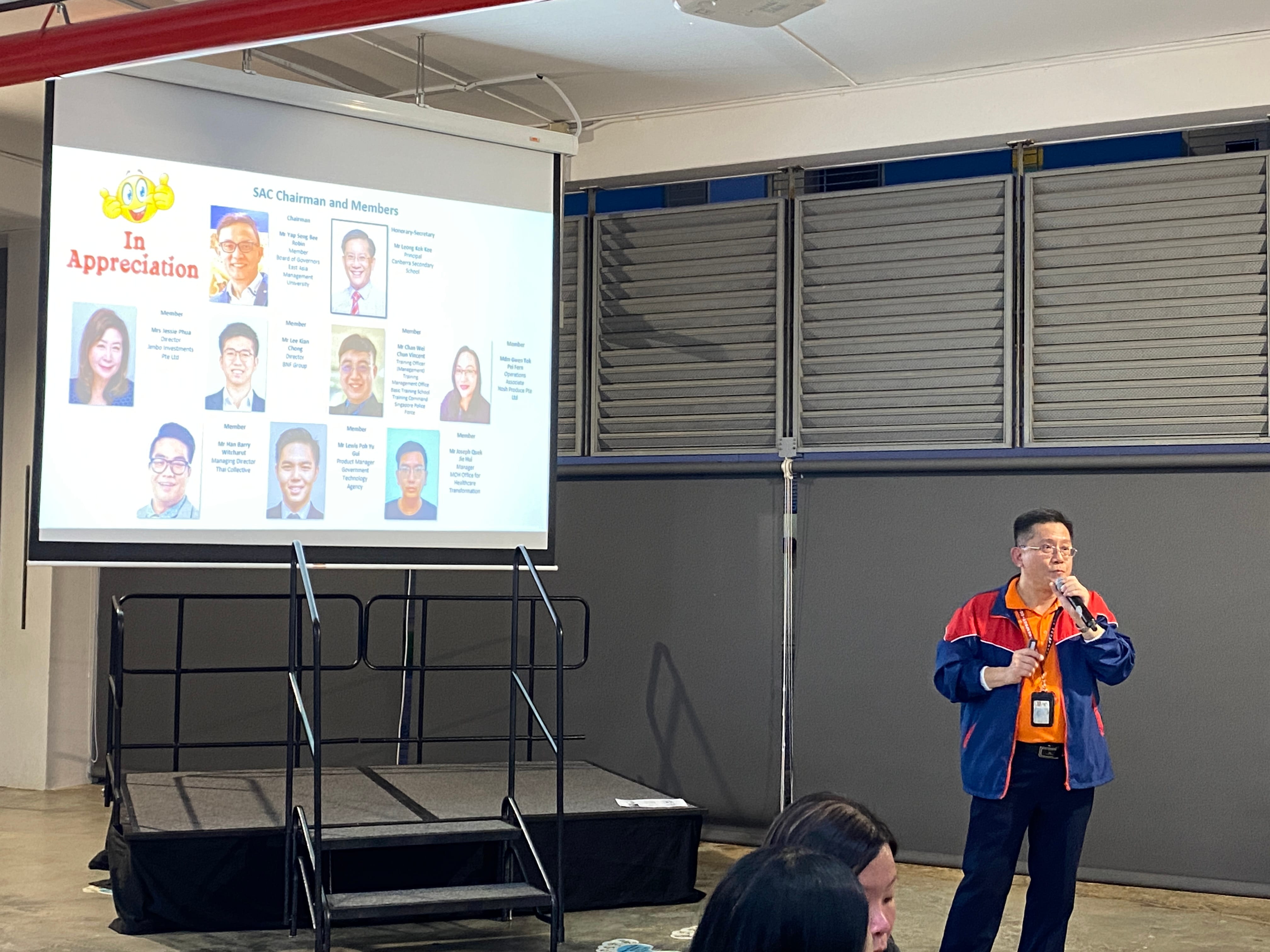 A man in a blue and red jacket speaks into a microphone beside a projected slide titled "In Appreciation" showing SAC Chairman and Members.