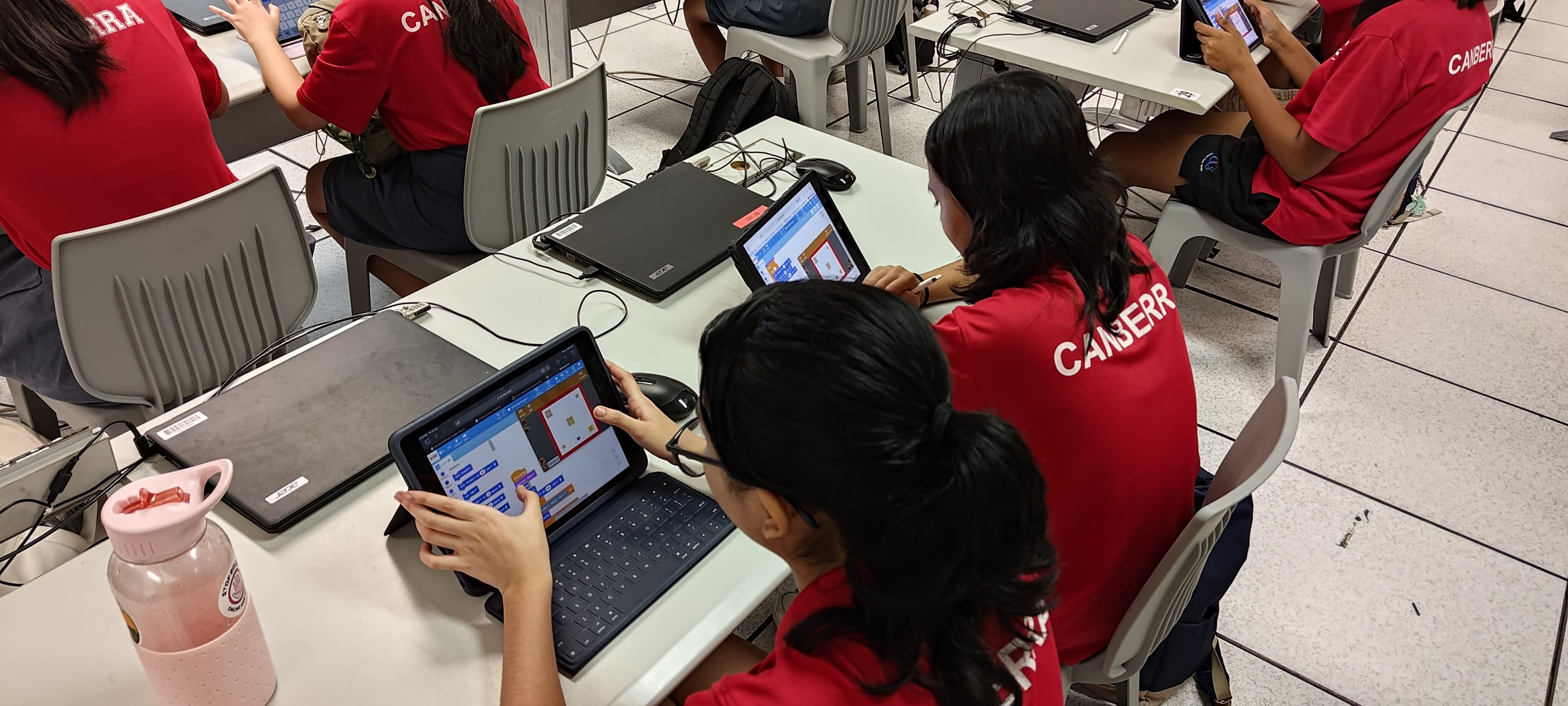Students in red Canberra shirts work on tablets with a coding interface in a classroom setting.