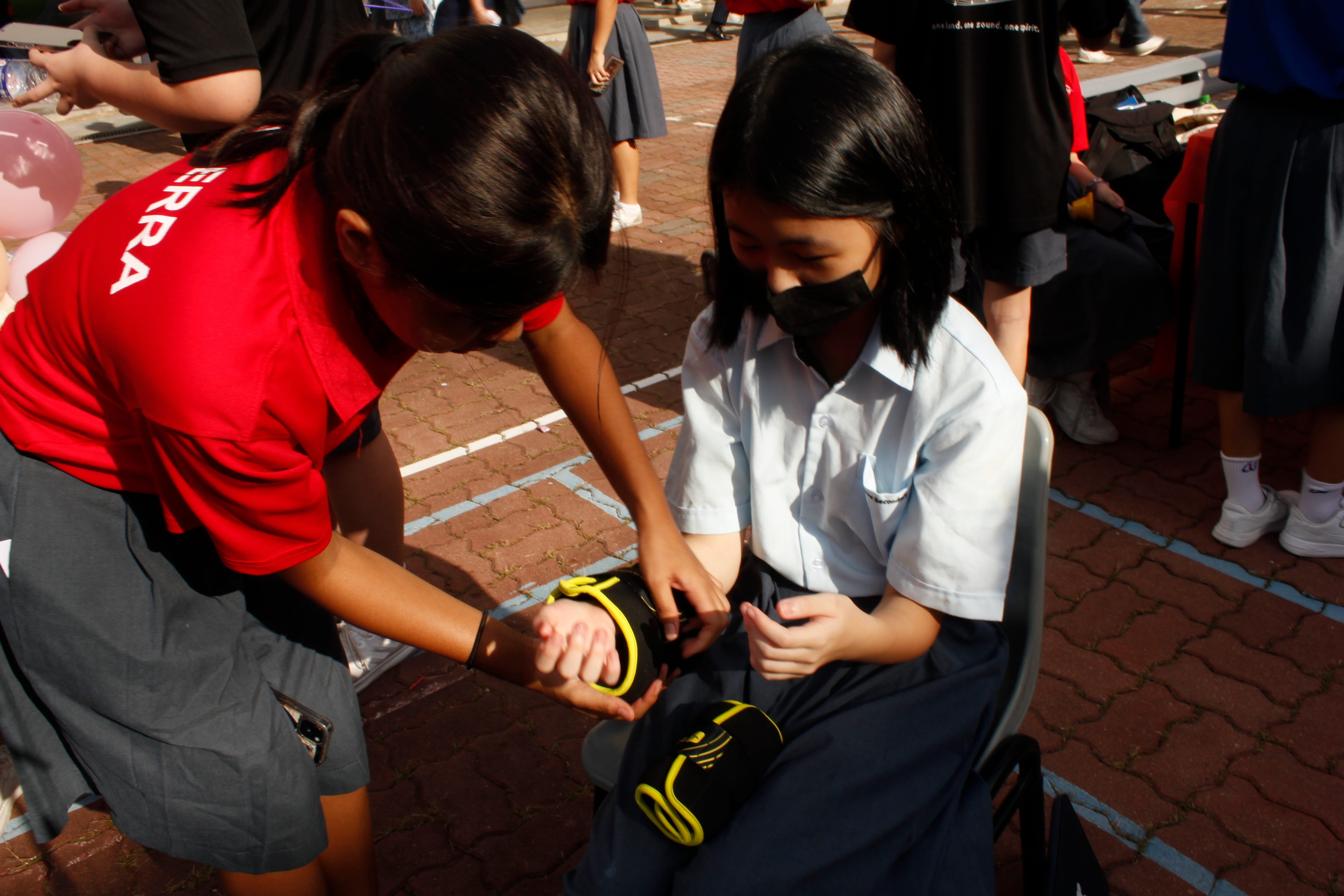 A student in a red tee puts on wrist weights on a student in a school uniform