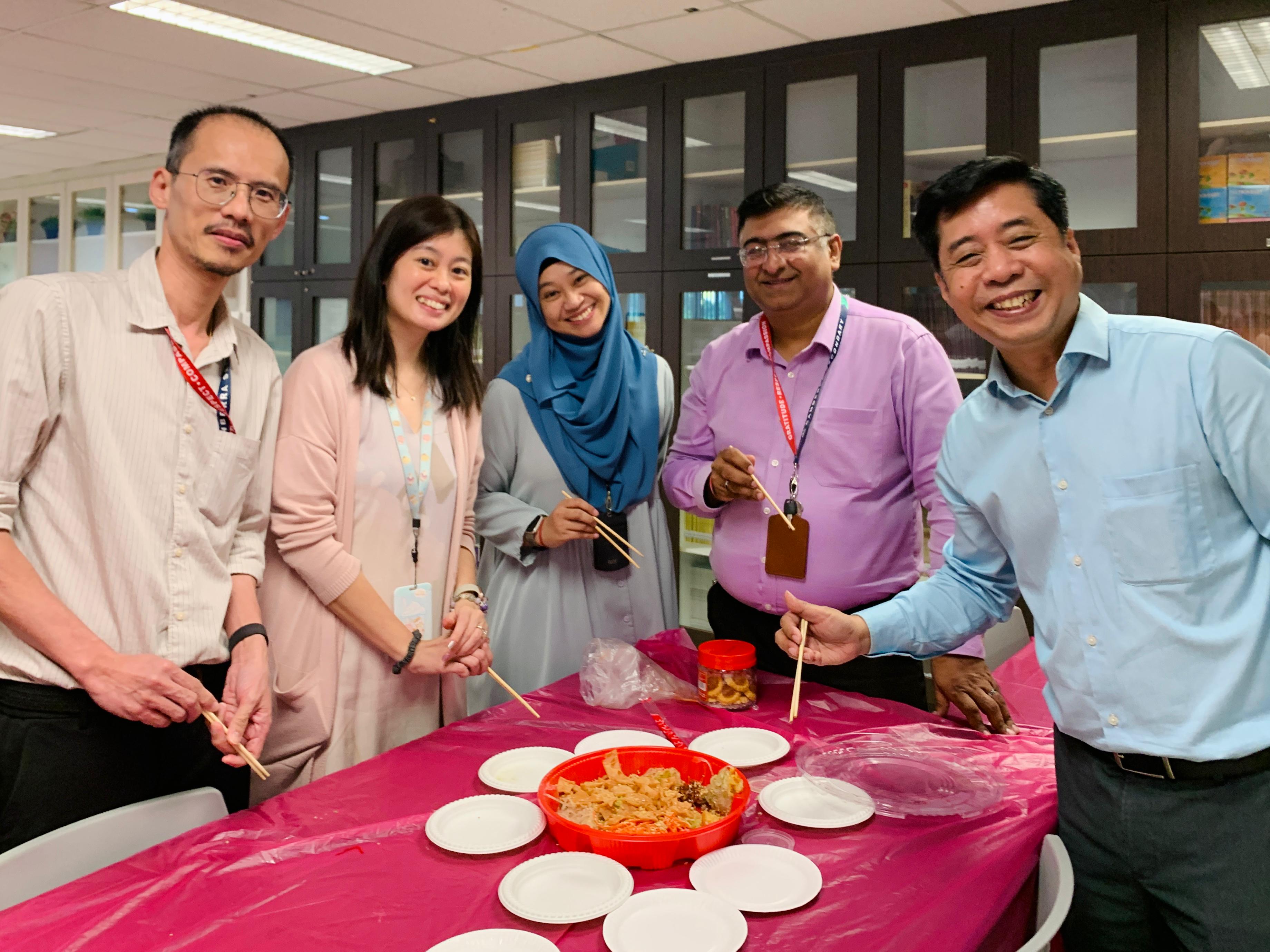 Five smiling colleagues hold chopsticks around a pink table with a red bowl of food and white plates.