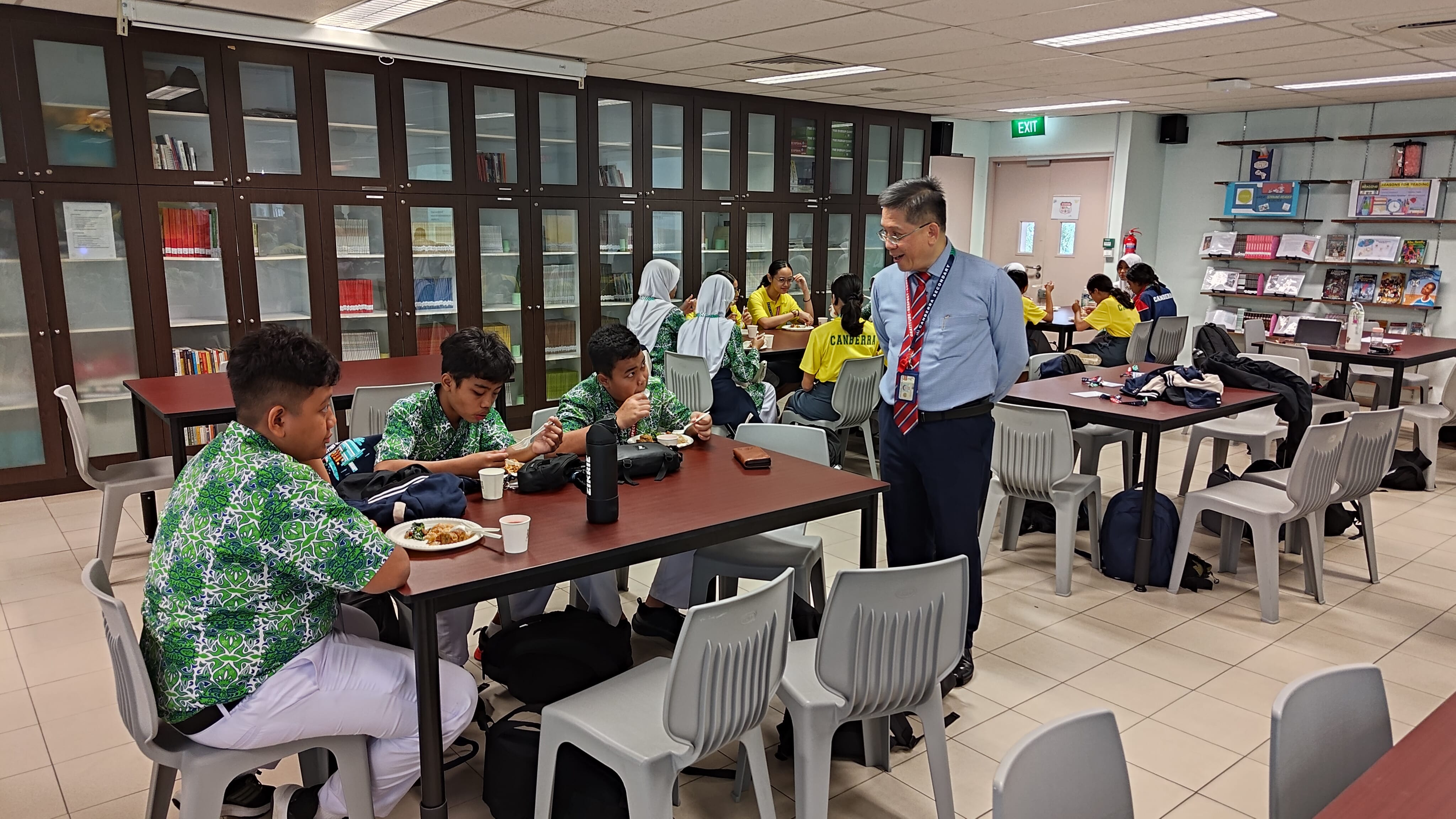 A teacher in a blue shirt and red tie speaks to students eating lunch in a school library with bookshelves.