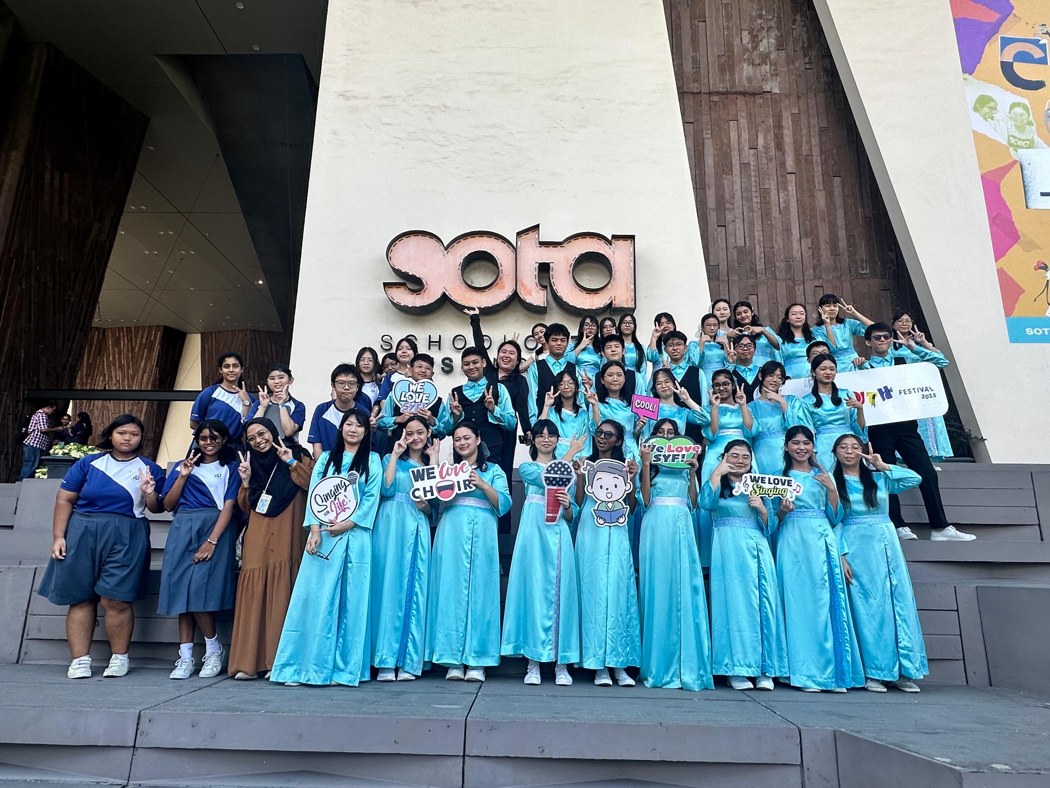 A large choir group in matching teal gowns poses on steps outside the SOTA building, holding signs about singing.