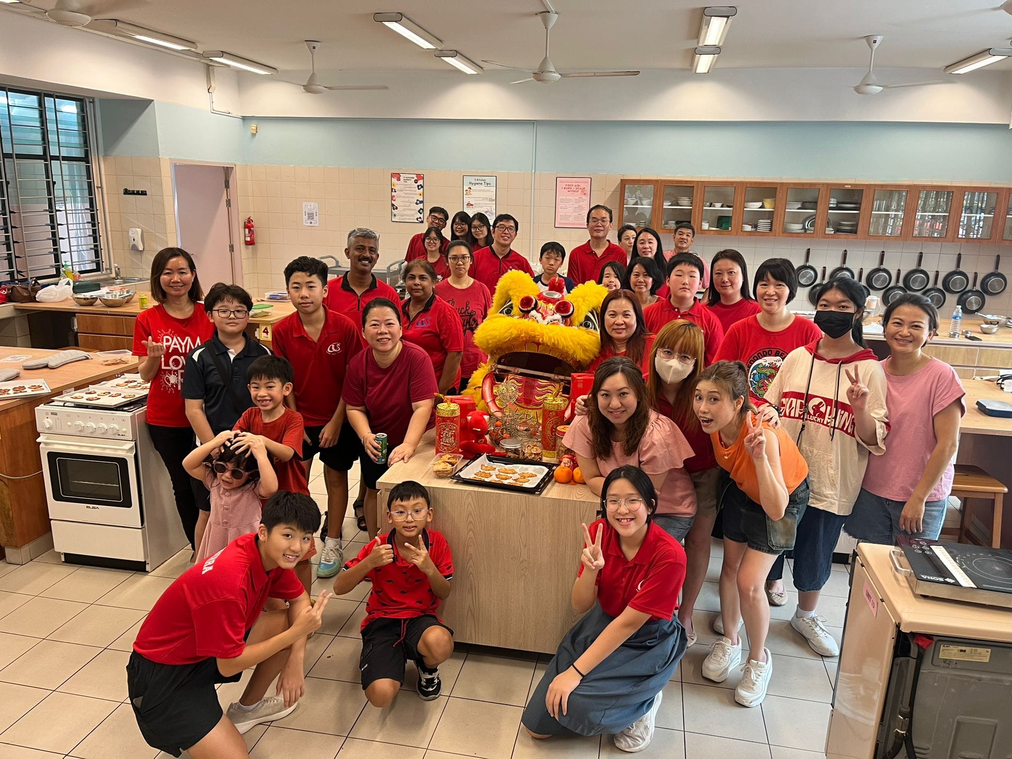 A large group in red shirts poses in a kitchen around a table with a lion dance head and festive decorations.