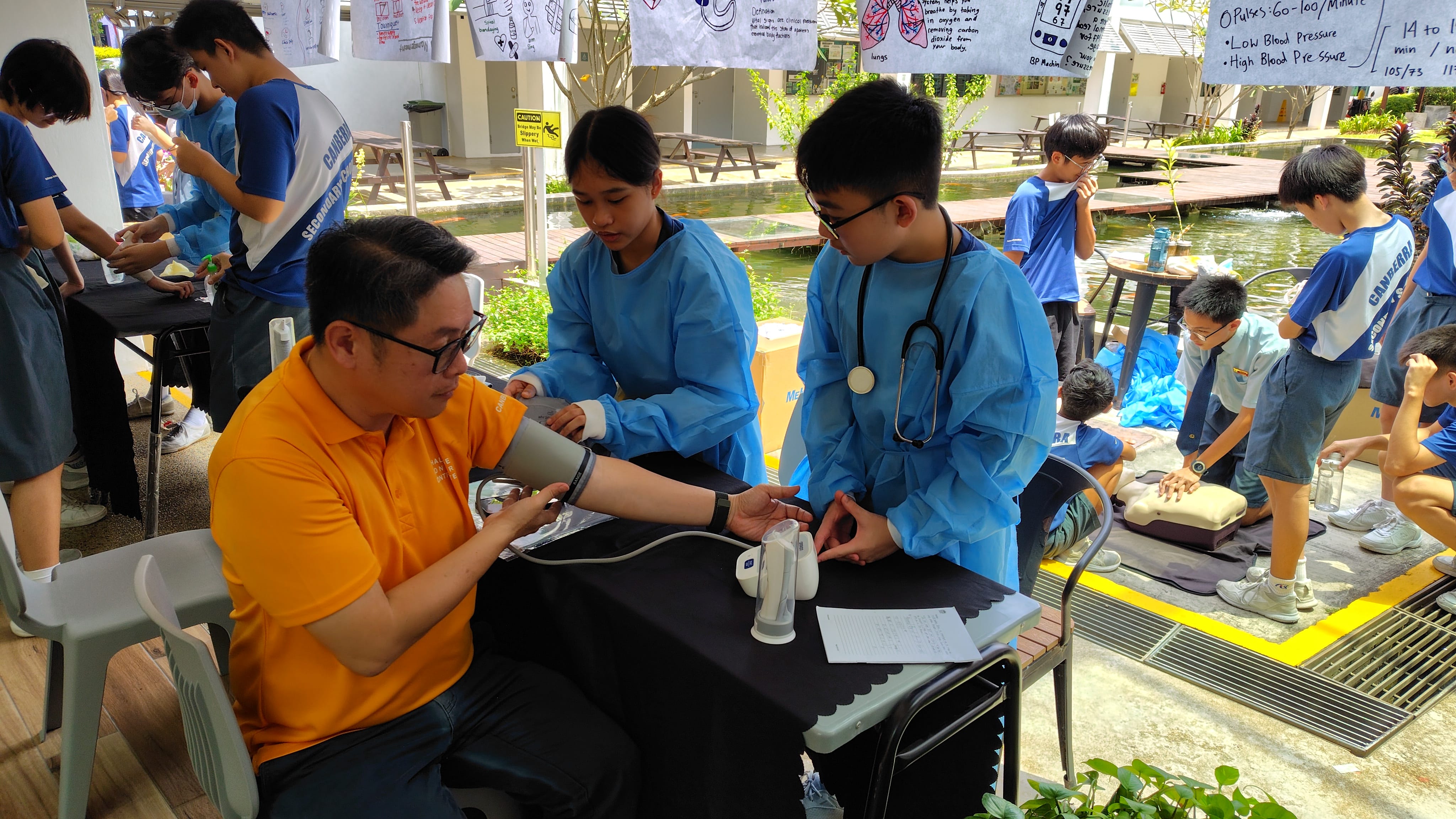 Two students in blue gowns take a man's blood pressure outdoors while others practice CPR on mannequins nearby.