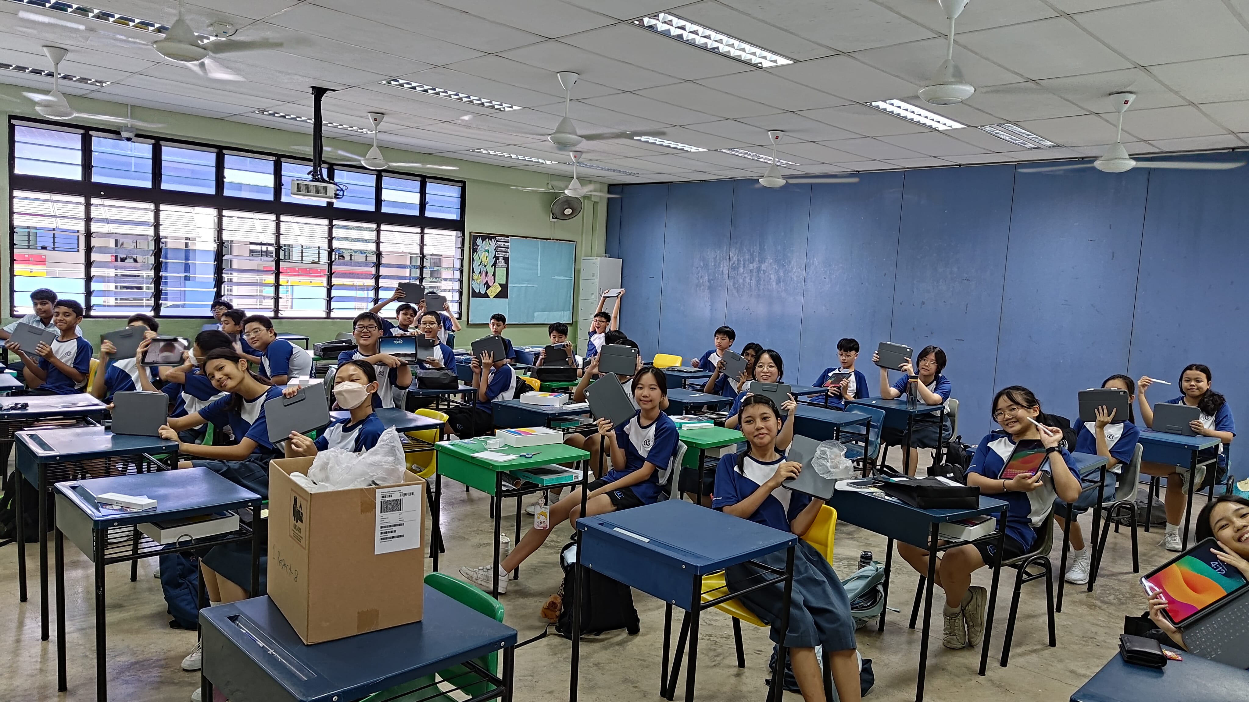 Students in blue uniforms sit at desks in a classroom, smiling and holding up tablets toward the camera.