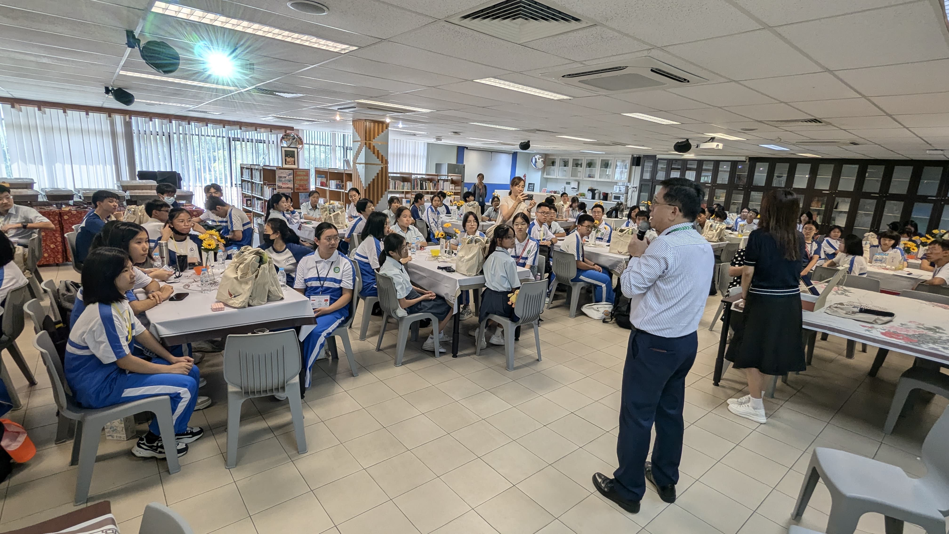 A man in a white shirt addresses a large group of students in blue and white uniforms seated at tables indoors.