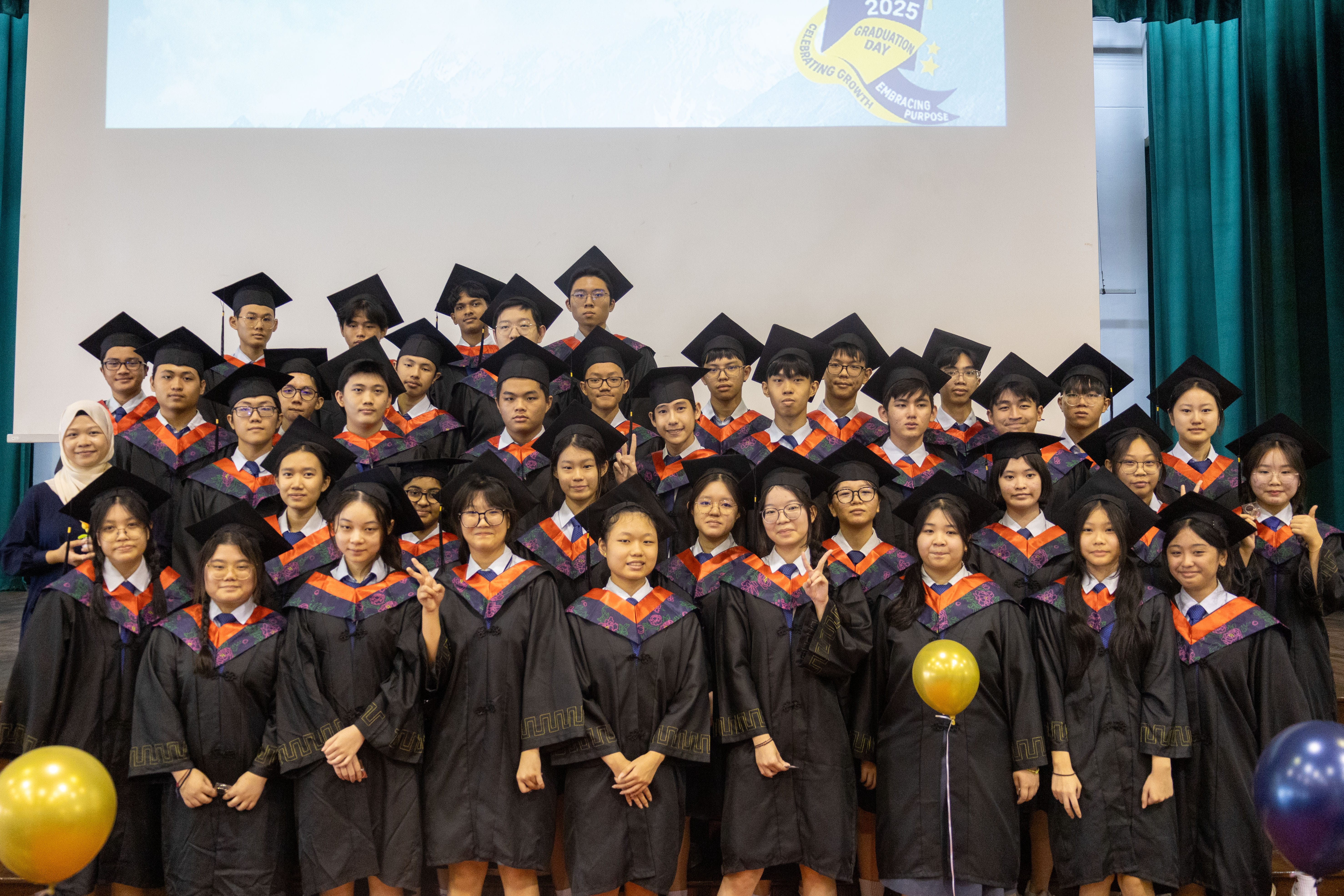 Group of graduates in caps and gowns posing on stage, some flashing peace signs, with balloons in front.
