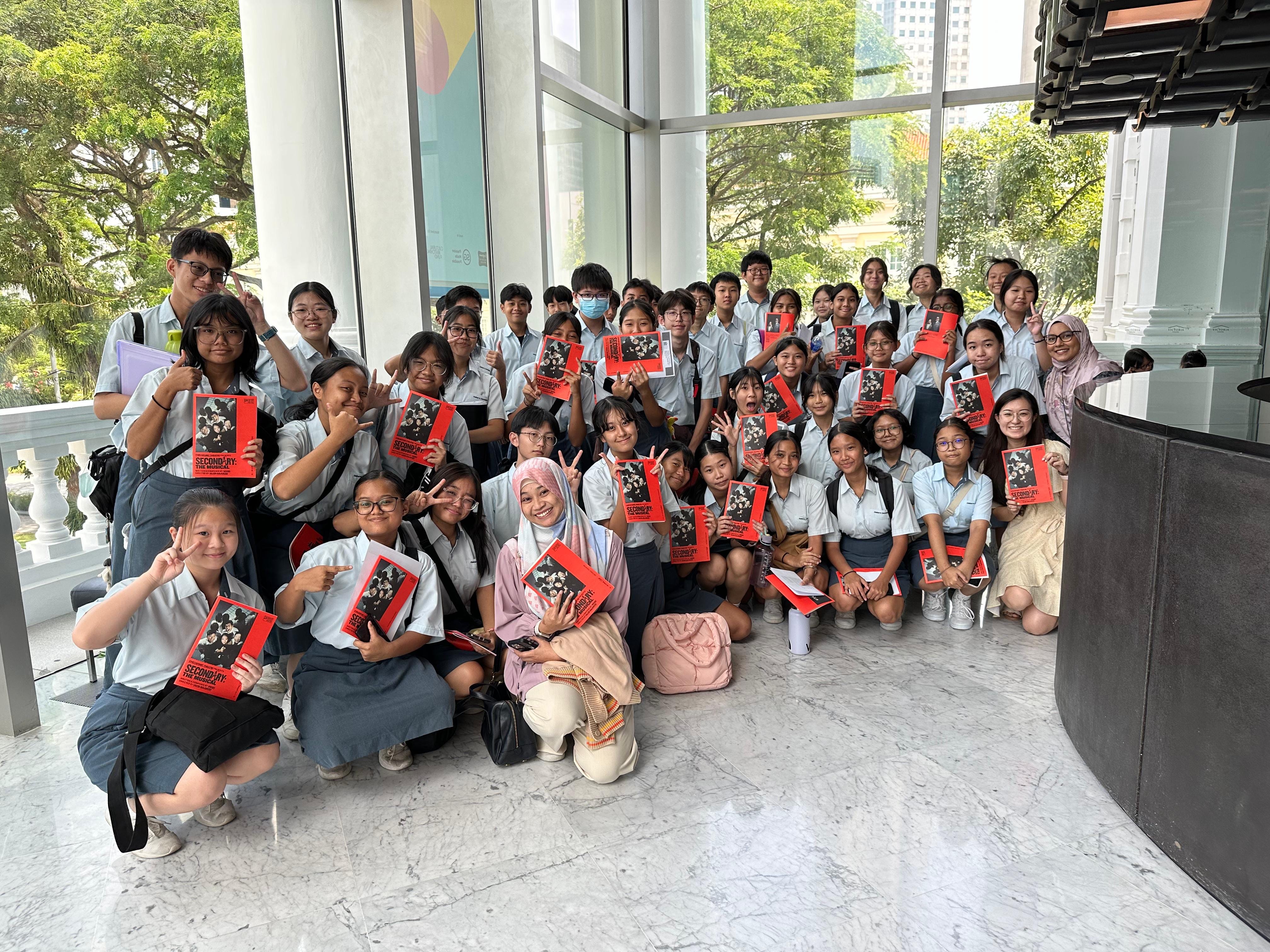 A large group of students in light blue uniforms posing indoors, holding red program booklets, smiling and making hand gestures.