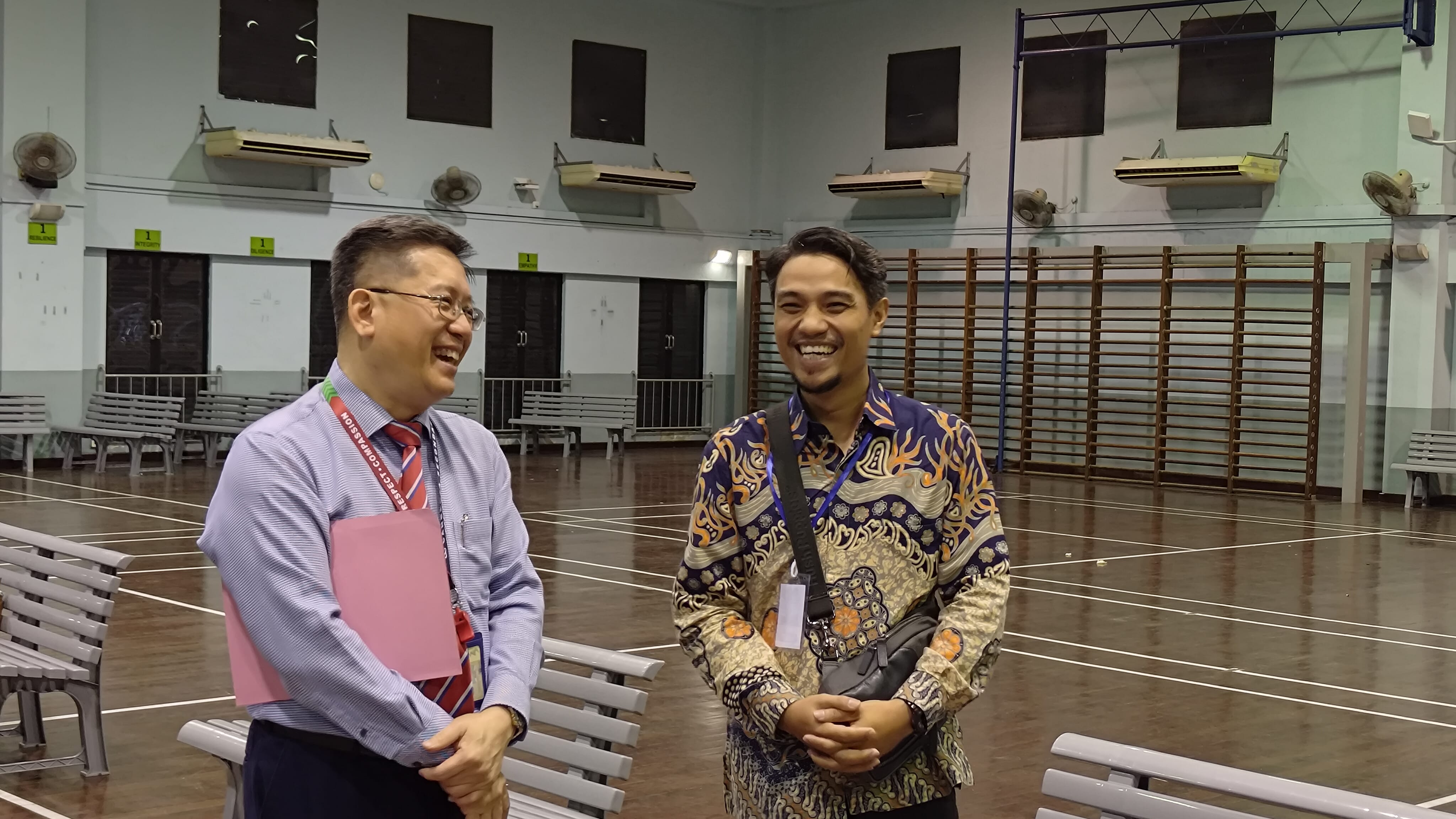 Two smiling men stand in a large indoor hall with benches, wall bars, and fans mounted on the walls.