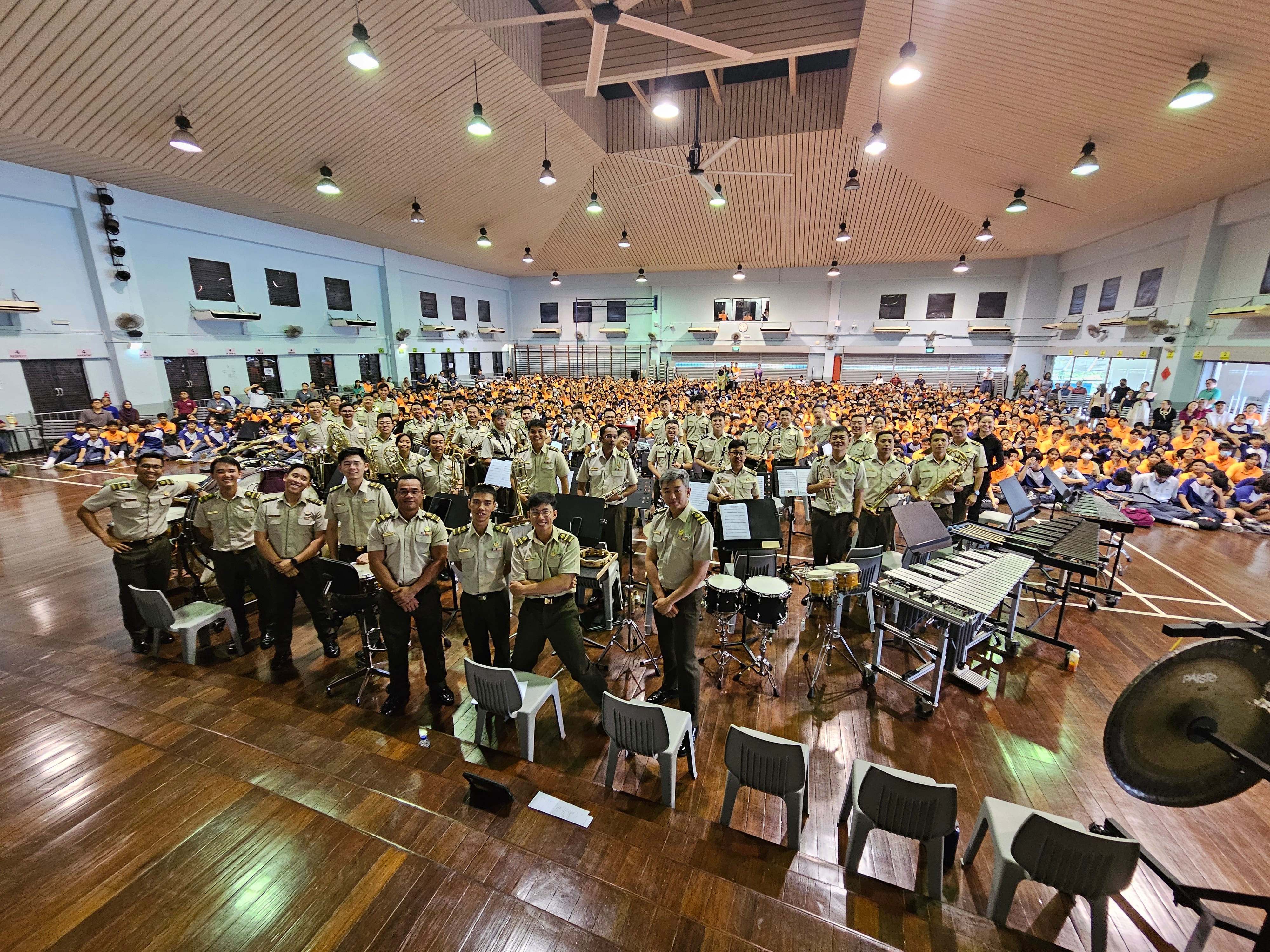 Uniformed band members pose by drums and xylophones in a gym hall packed with seated students.