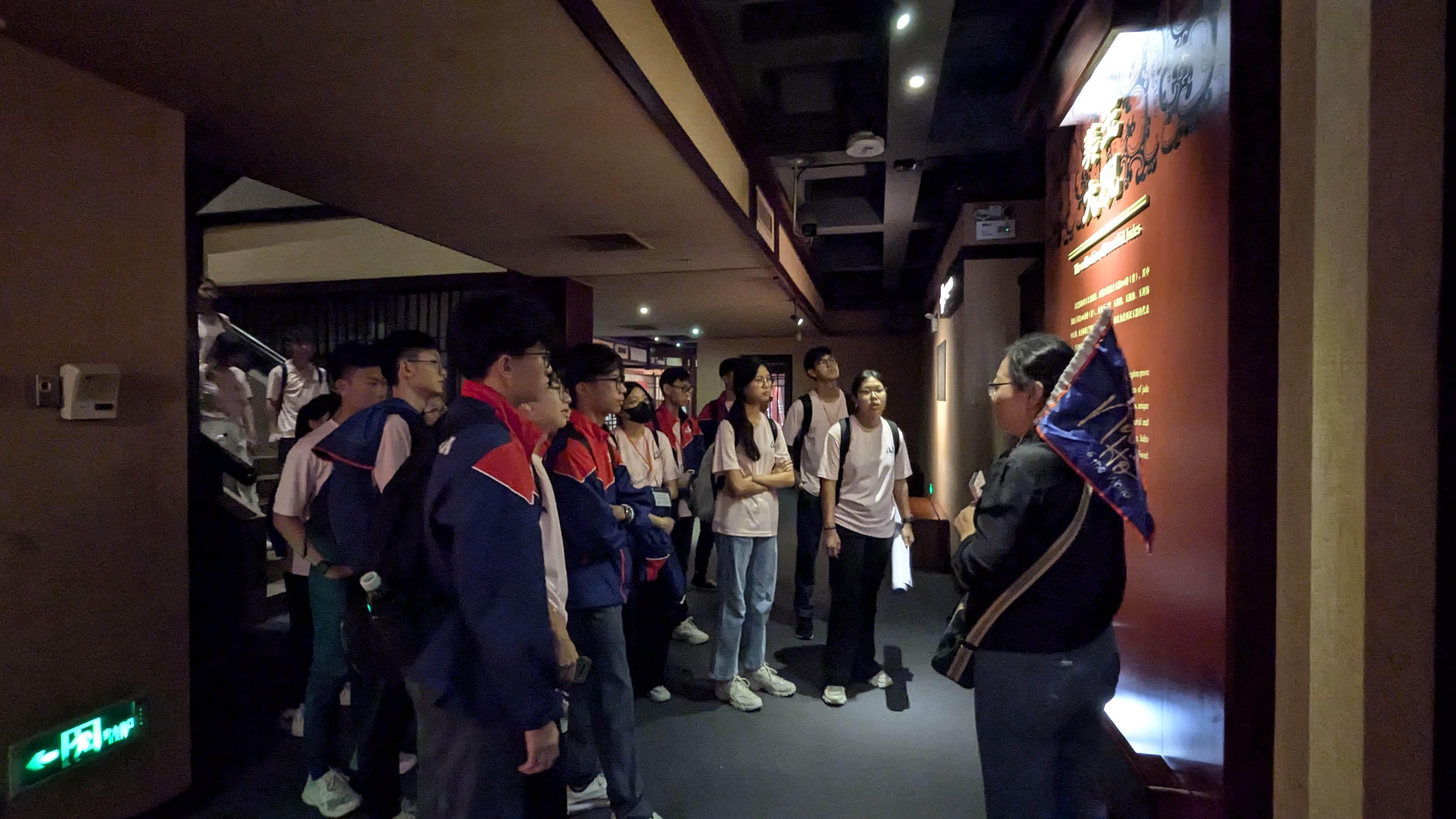 A tour guide leads a group of young students through a dimly lit museum hallway with Chinese text on red walls.