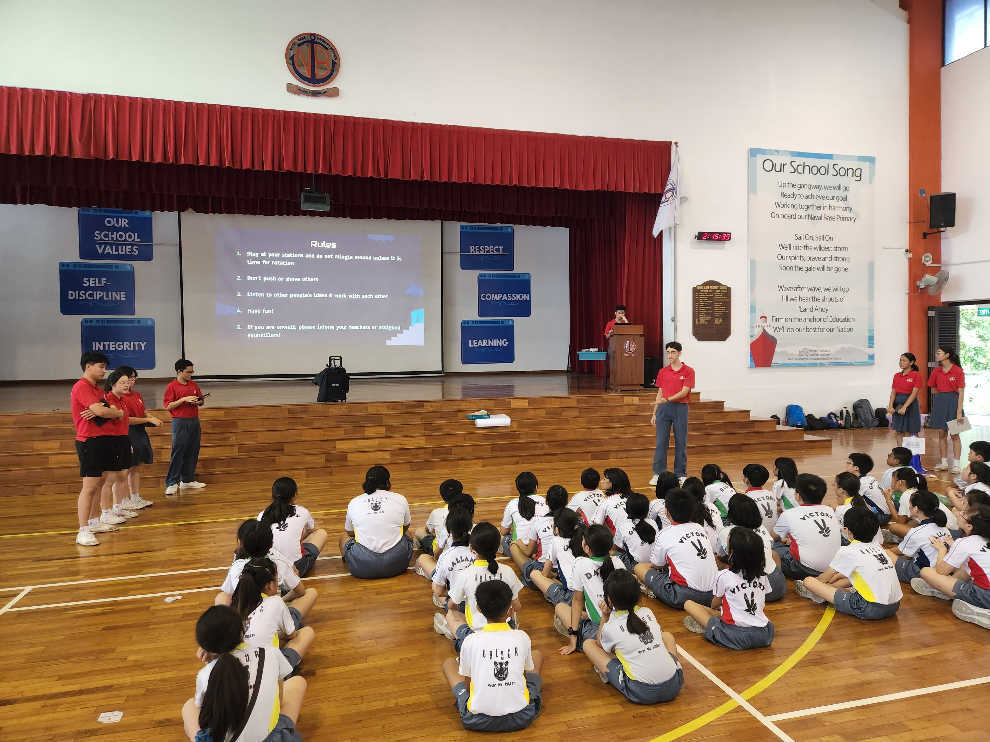Students in red tees address a group of primary school students in their school hall