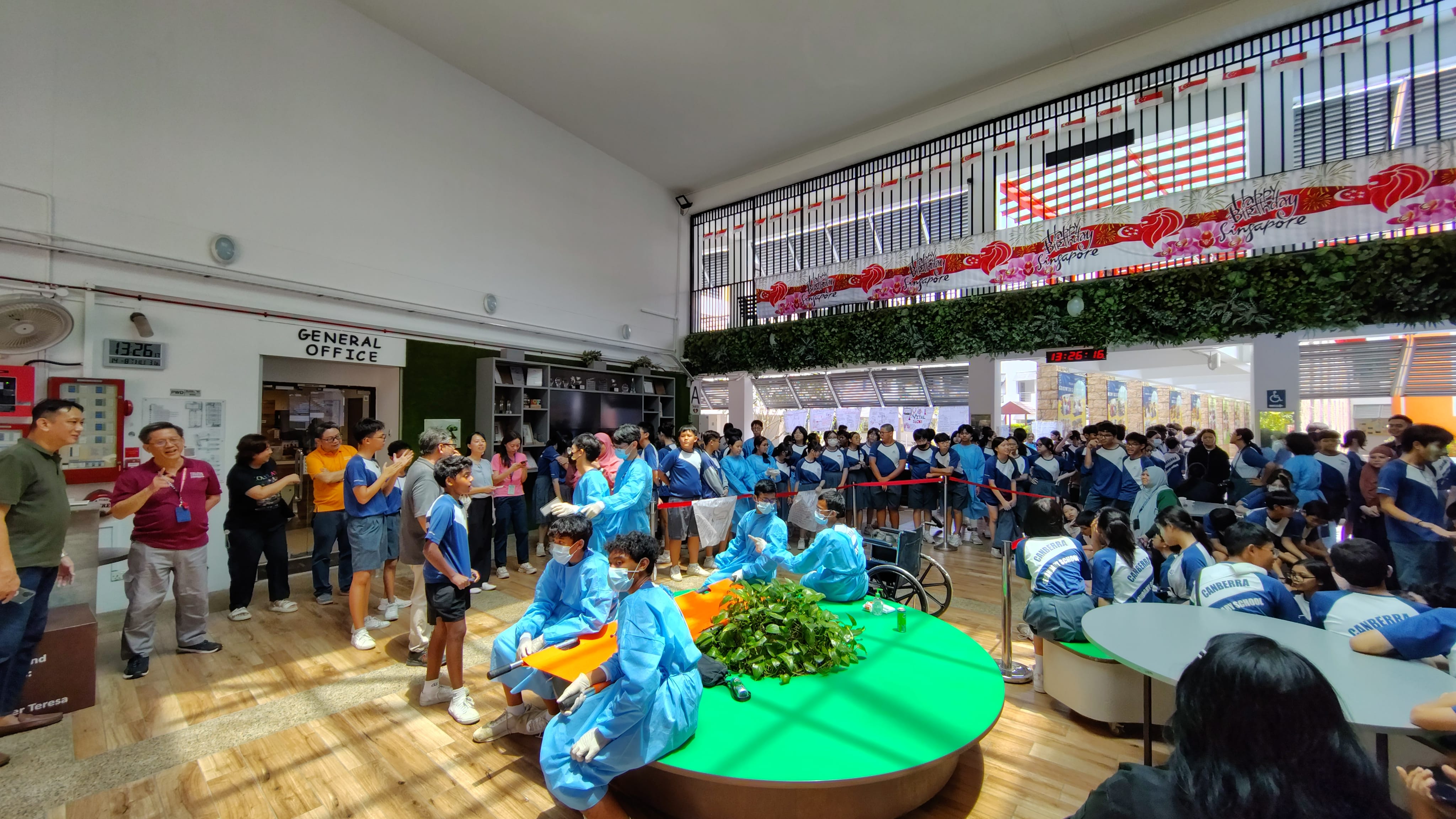 Students in blue medical gowns and masks perform a first aid drill watched by a crowd in a school atrium.