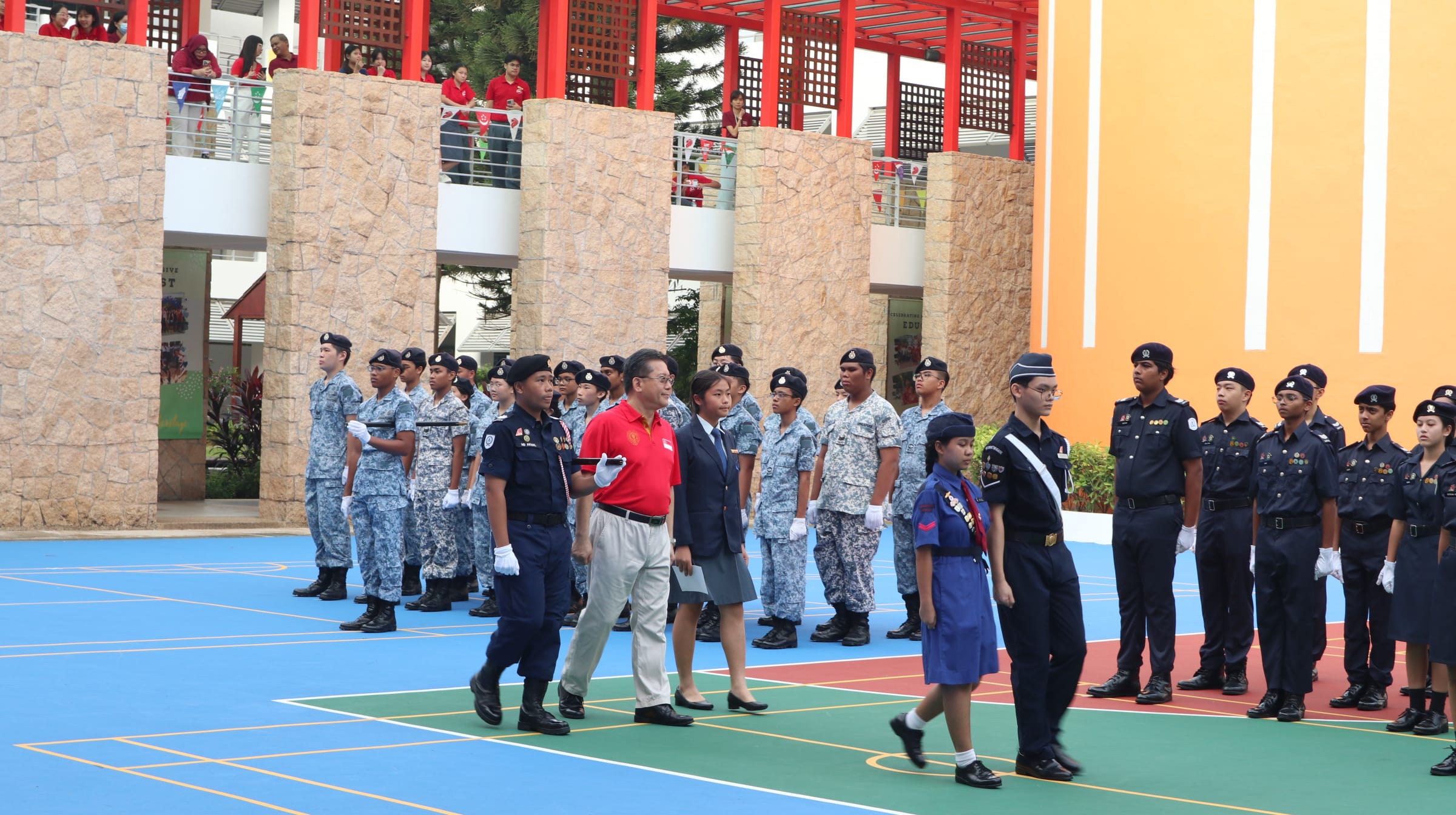 Students in military and scout uniforms stand at attention on a colorful court during a school parade ceremony.
