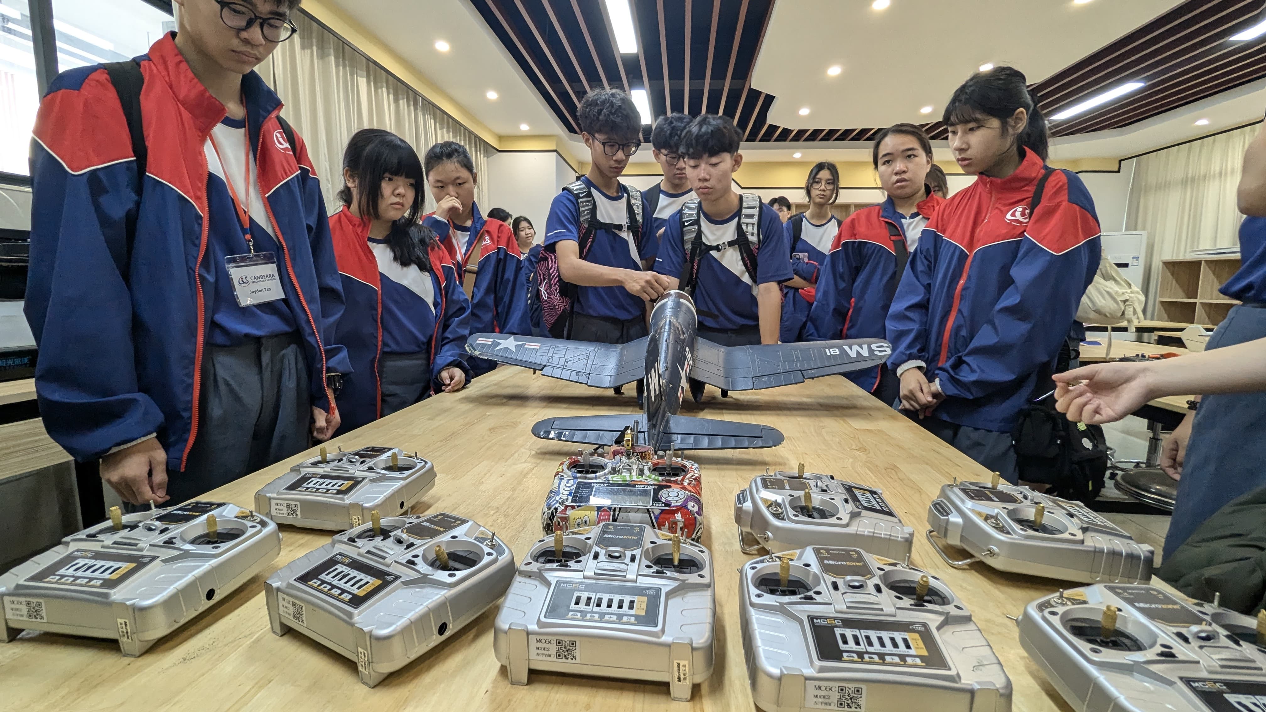 Students in blue and red jackets examine an RC airplane and multiple radio controllers on a wooden table.