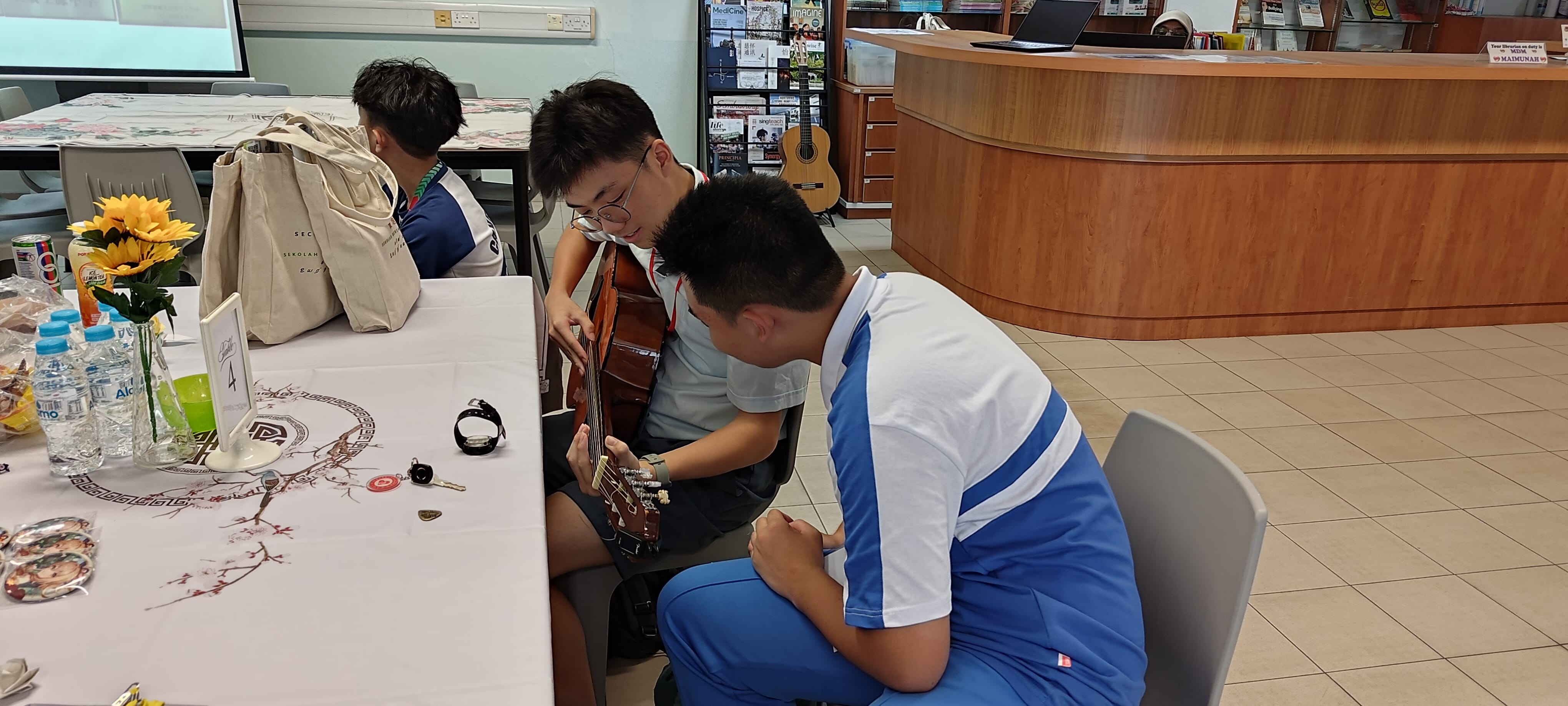 Two teenage boys sit together, one playing an acoustic guitar while the other watches closely, inside a library.