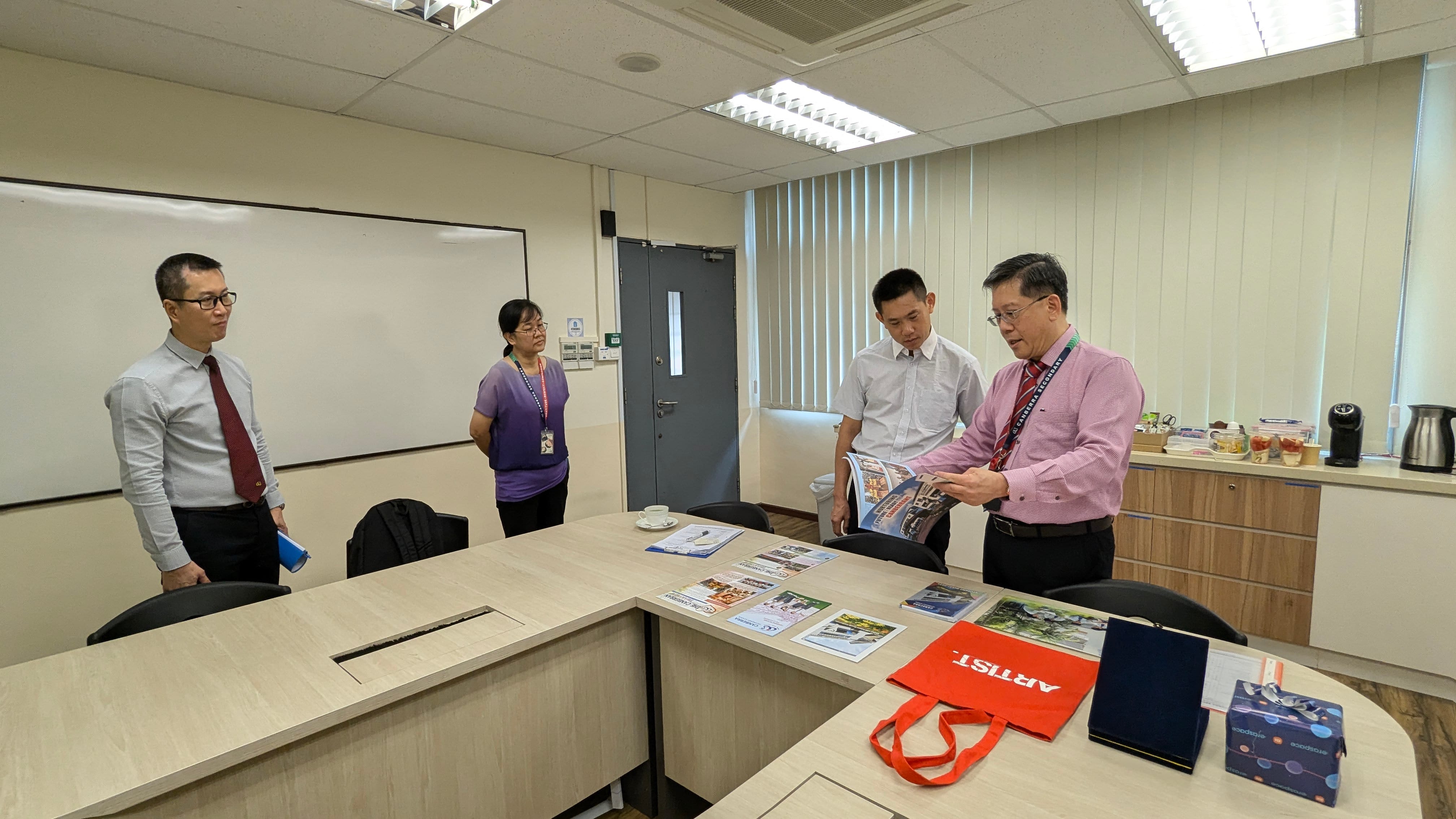 Four people stand in an office meeting room with brochures and a red bag on a light wood table.