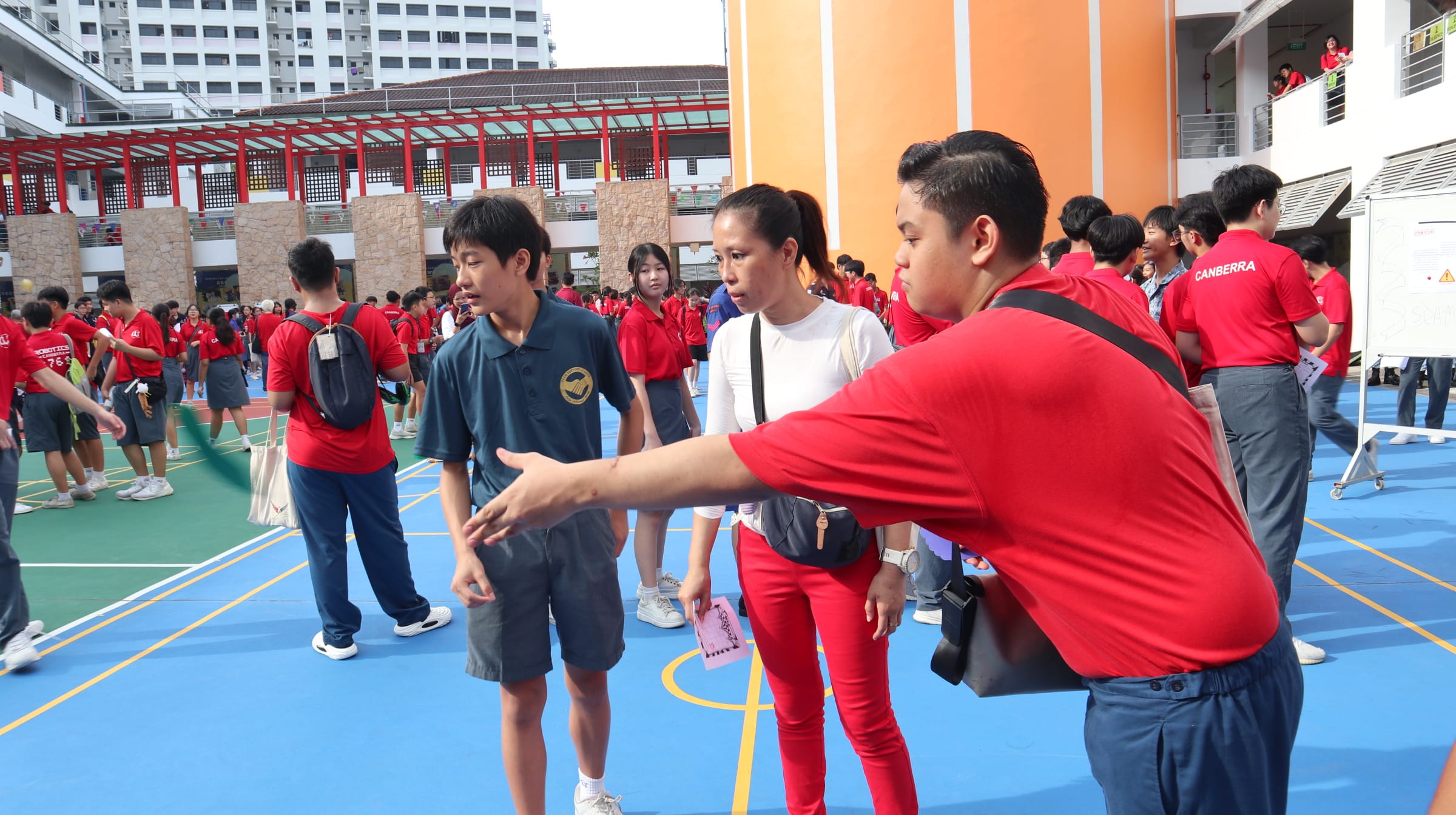 A student in red points directions to a boy and woman on a blue school courtyard surrounded by students in red uniforms.