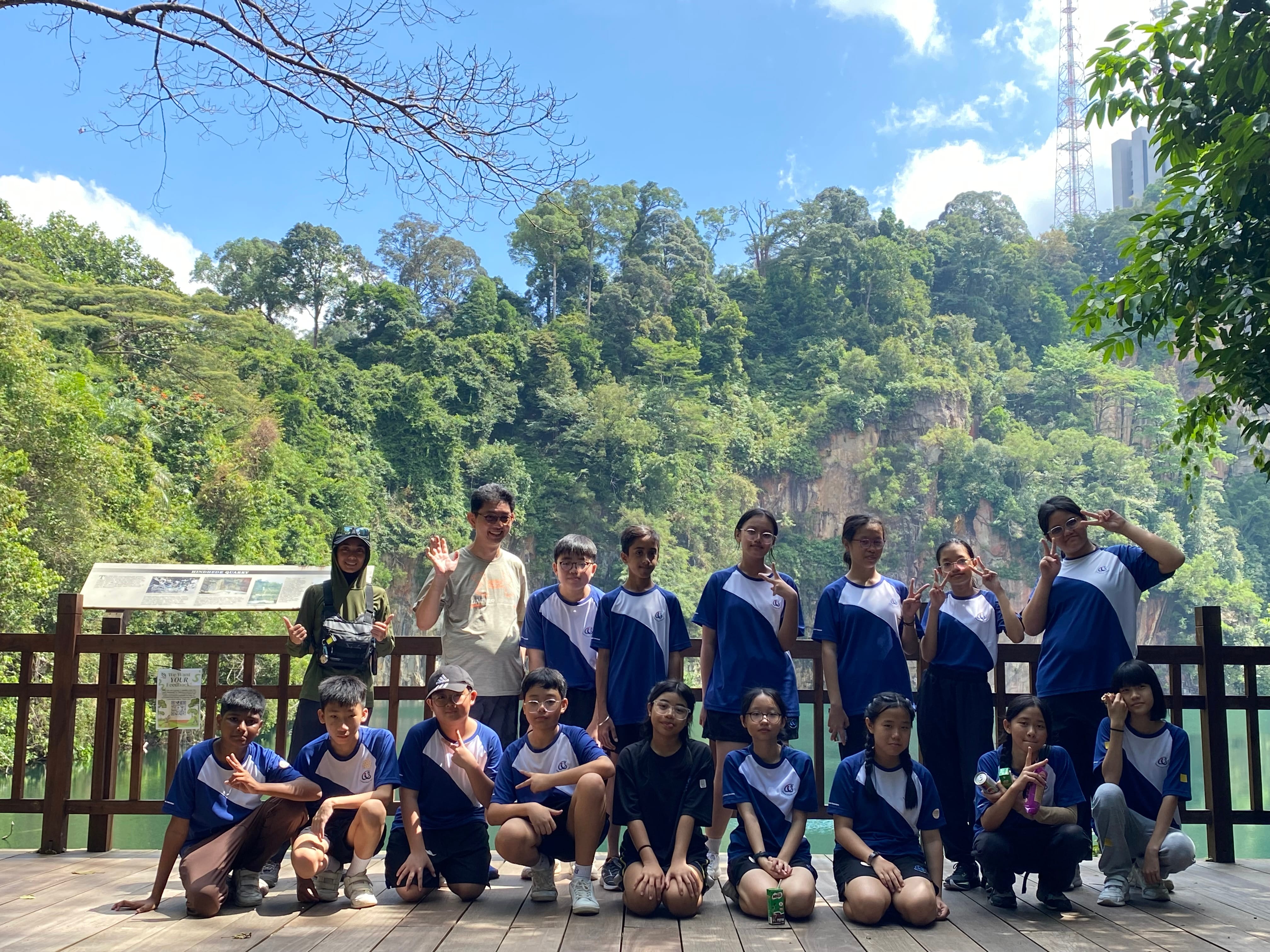 Group of students in blue uniforms posing on a deck, with lush forested cliffs and blue sky behind.