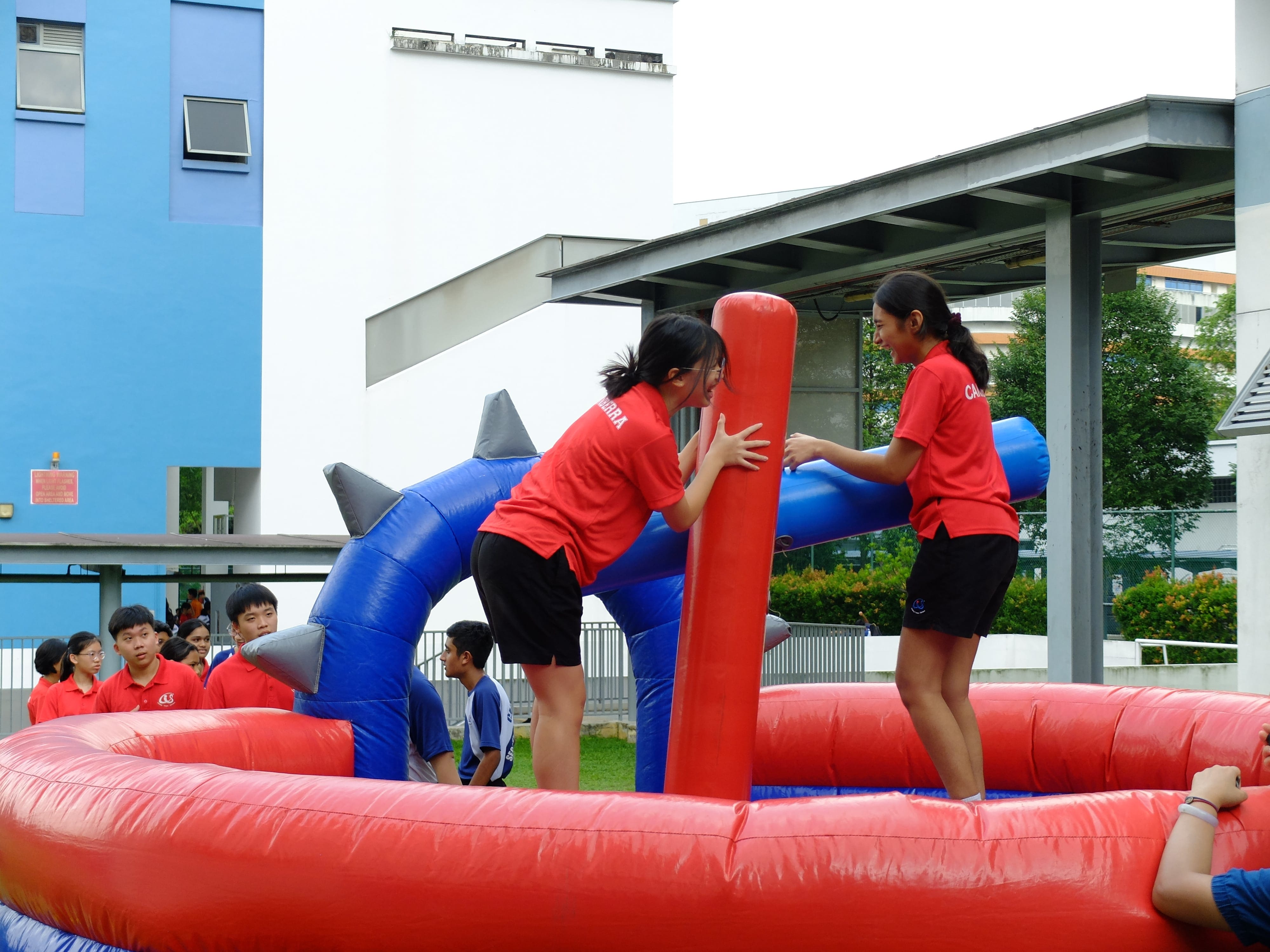Two students in red shirts play on a red and blue inflatable obstacle course outdoors near a building.
