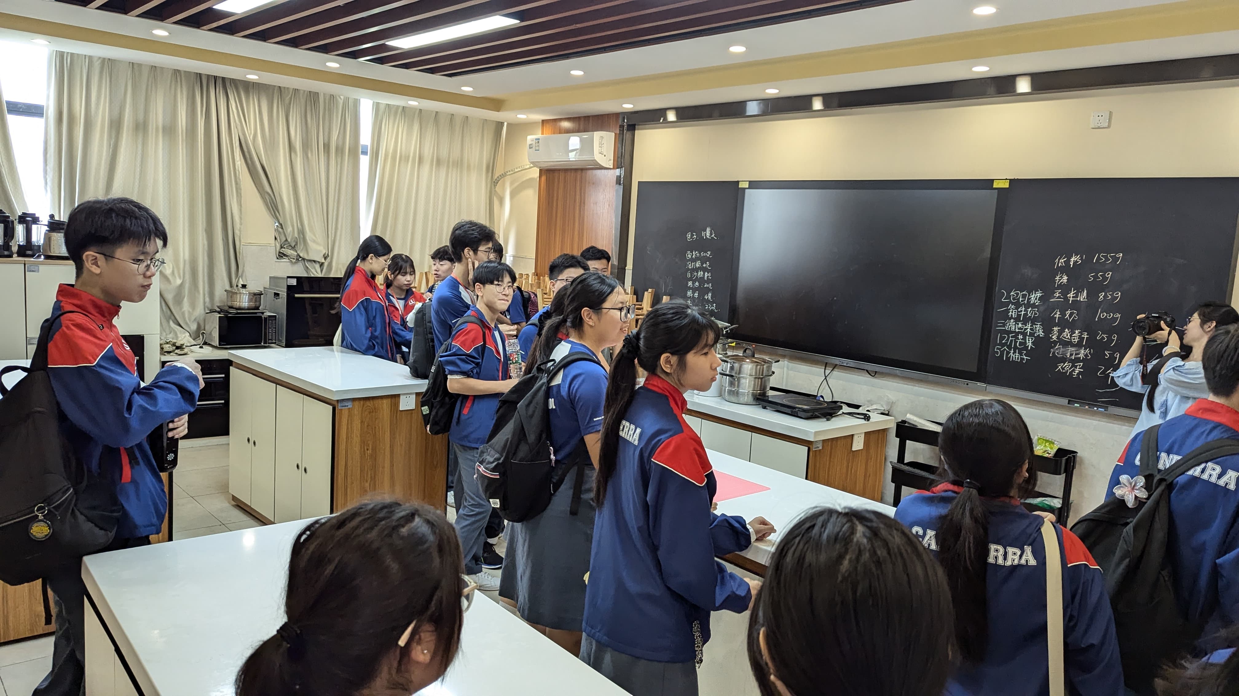 Students in blue and red uniforms stand in a classroom with a chalkboard showing Chinese writing and kitchen counters.