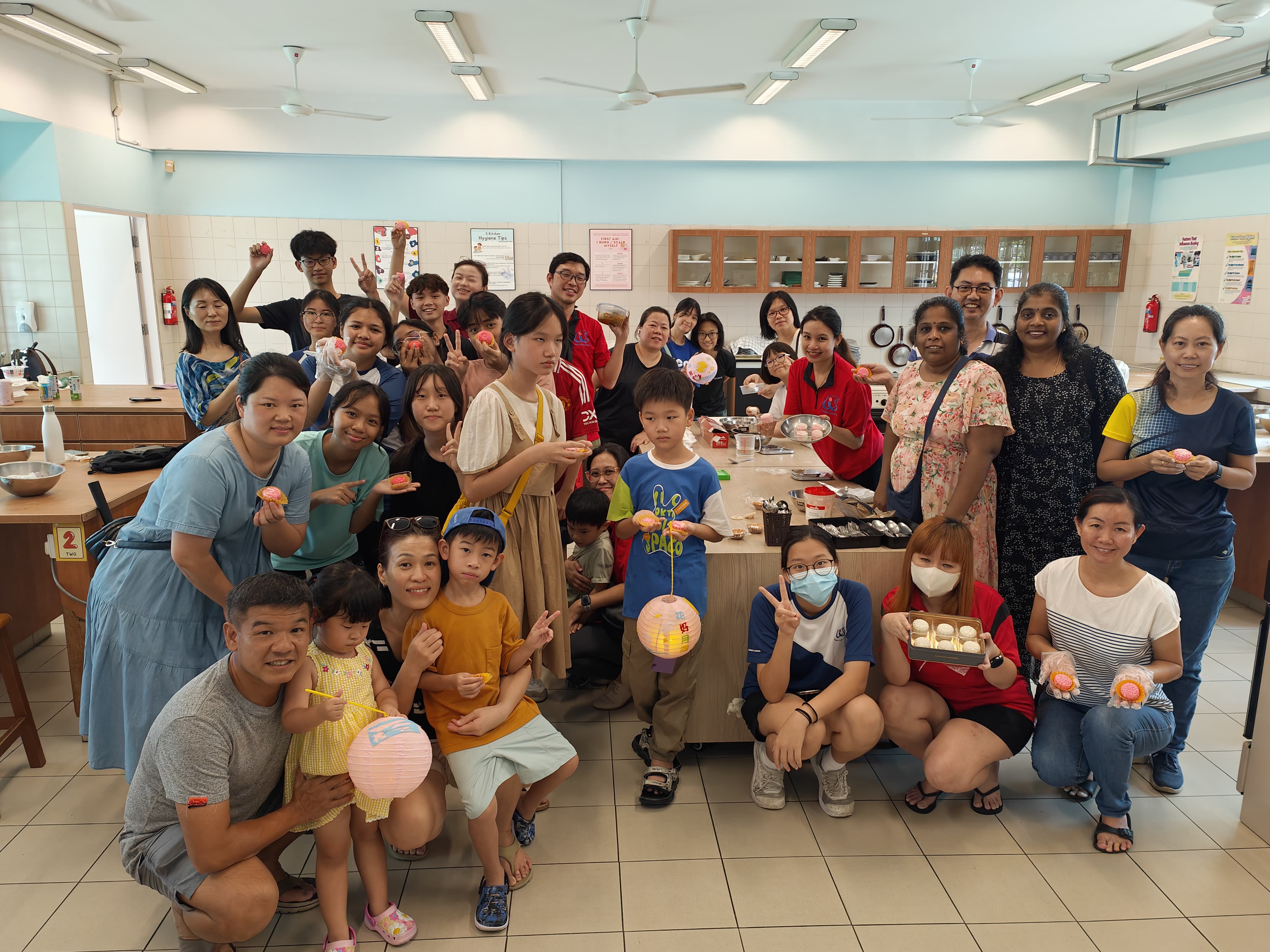 A large group of adults and children pose in a kitchen classroom holding mooncakes and paper lanterns.