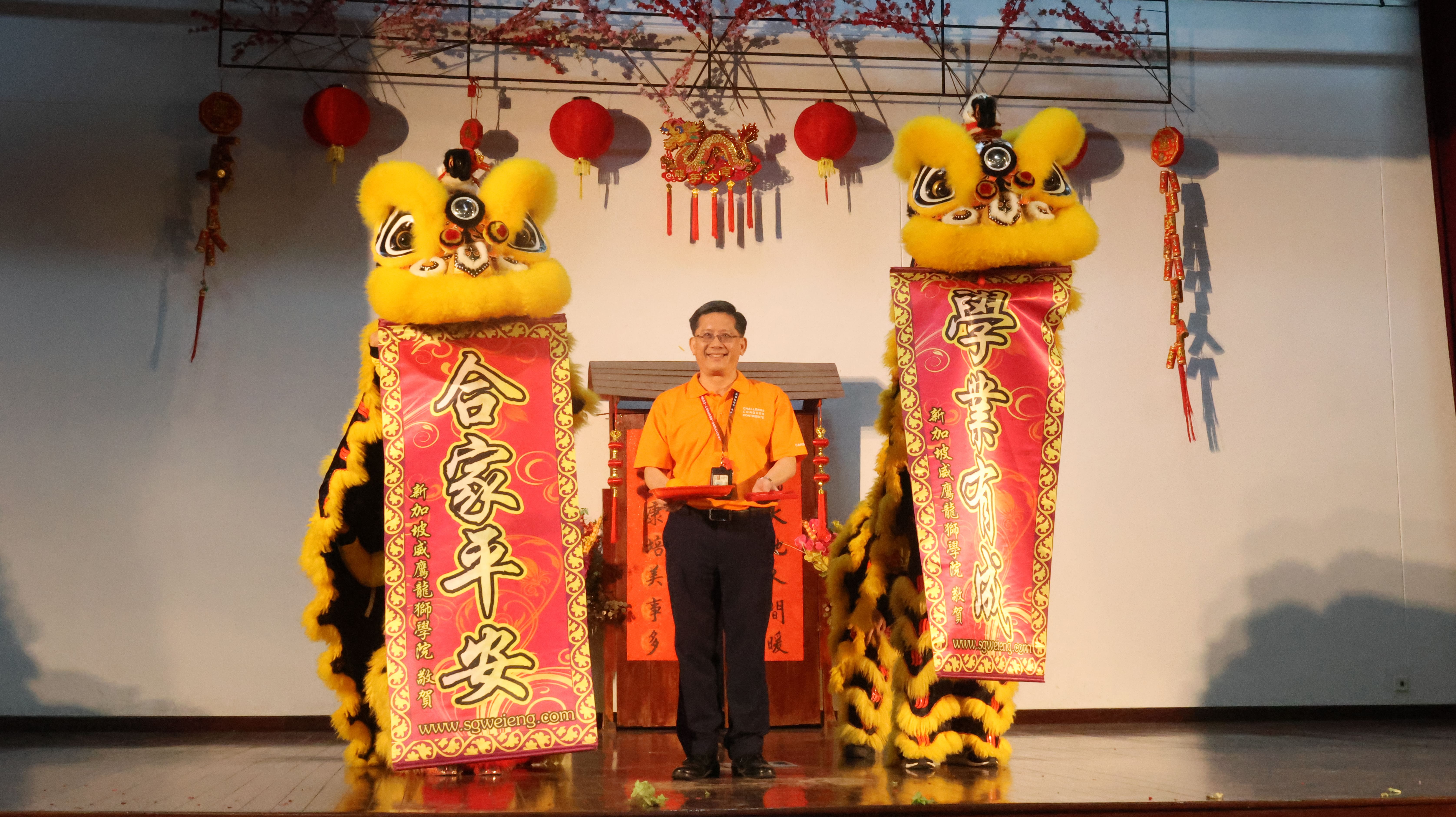 Man in orange shirt stands between two yellow Chinese lion dance costumes on a stage with red lanterns and banners.