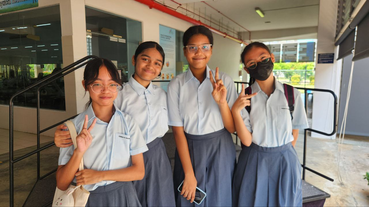 Four female students in light blue uniforms and gray skirts pose with peace signs outside a school building.