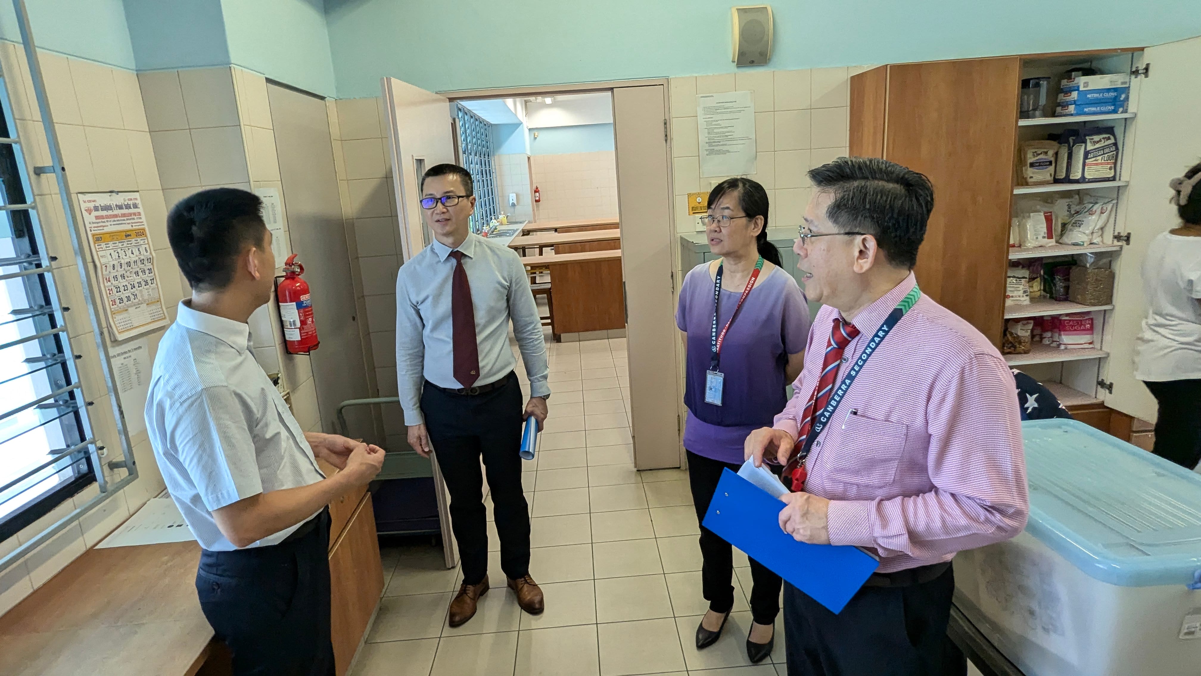 Four people in formal attire having a discussion inside a kitchen or utility room with shelves and a fire extinguisher.