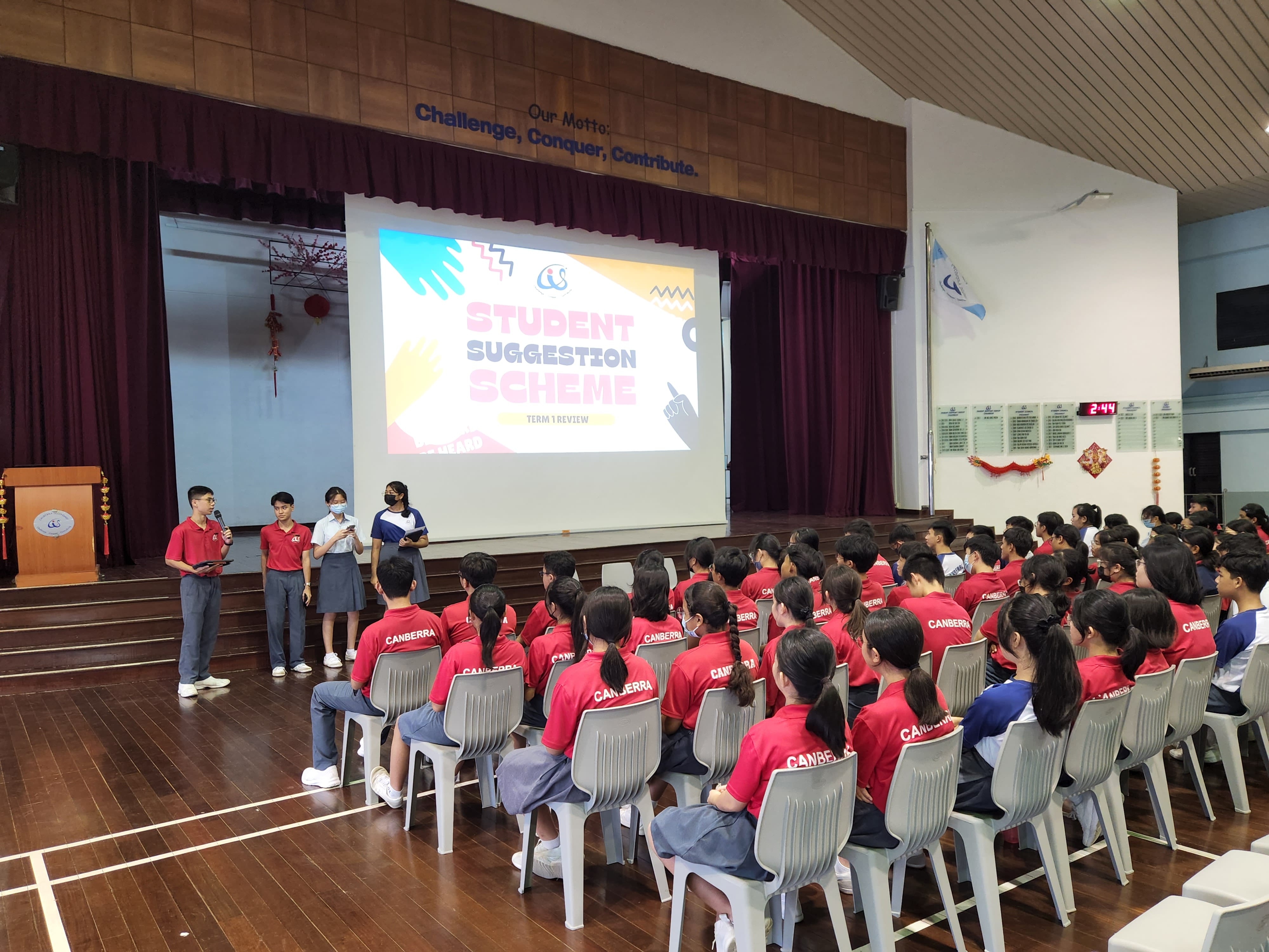 Four students at the front of the school hall adress the rest of the students seated on chairs, as a large screen shows the words 'Student Suggestion Scheme'