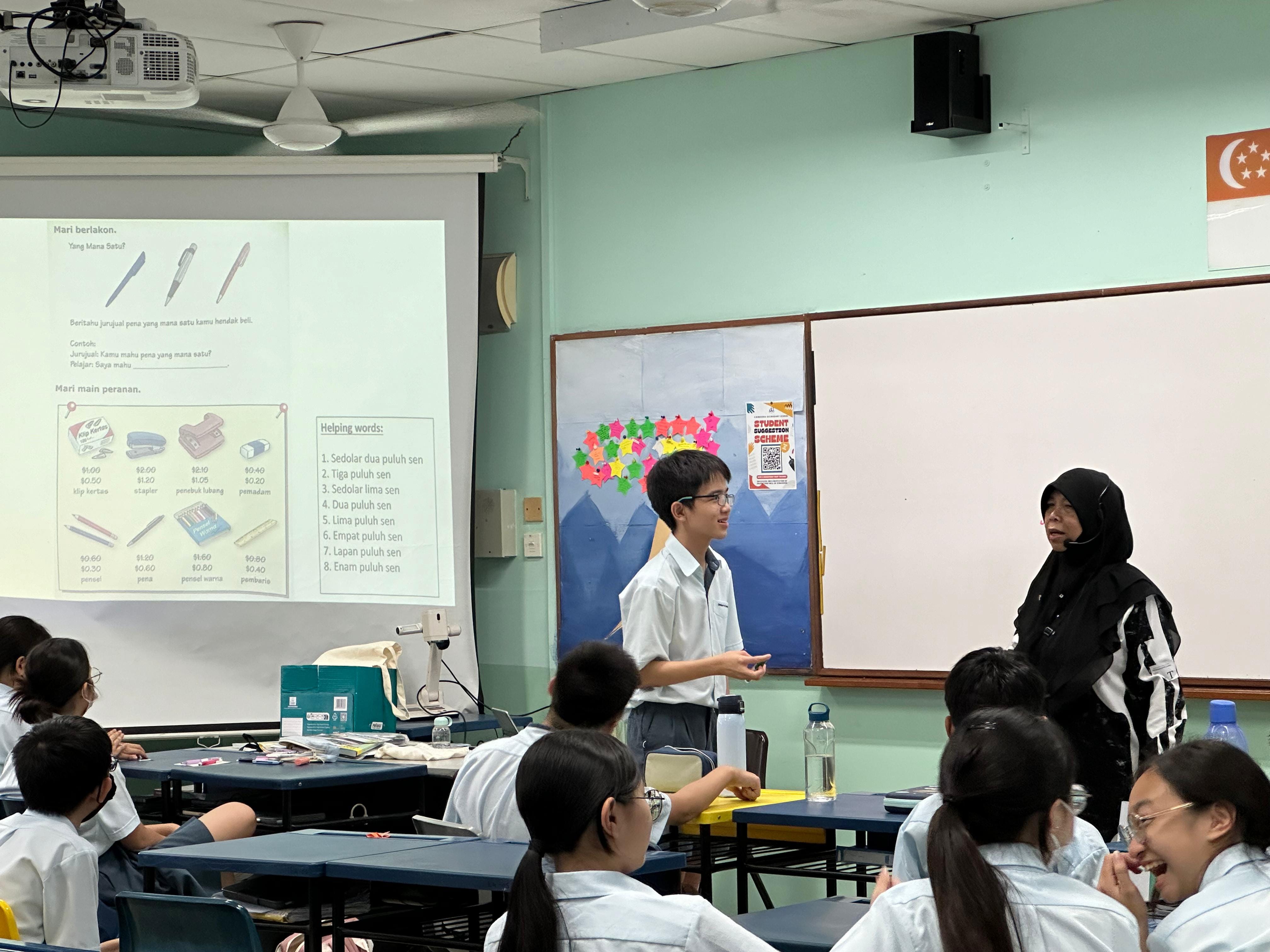 A student presents to classmates in a Singapore school classroom with a Malay language lesson projected on screen.