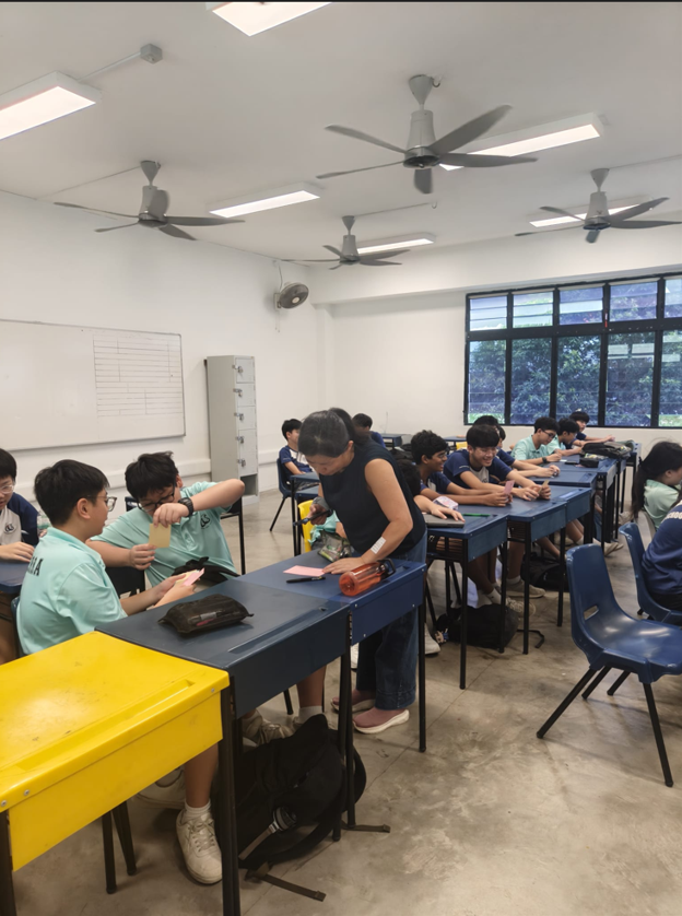 A teacher in a dark blue top moves between rows of desks to assist students during a classroom activity. Students in teal and navy Canberra Secondary School uniforms are seated at blue desks in a bright, modern classroom with large ceiling fans and windows overlooking greenery.