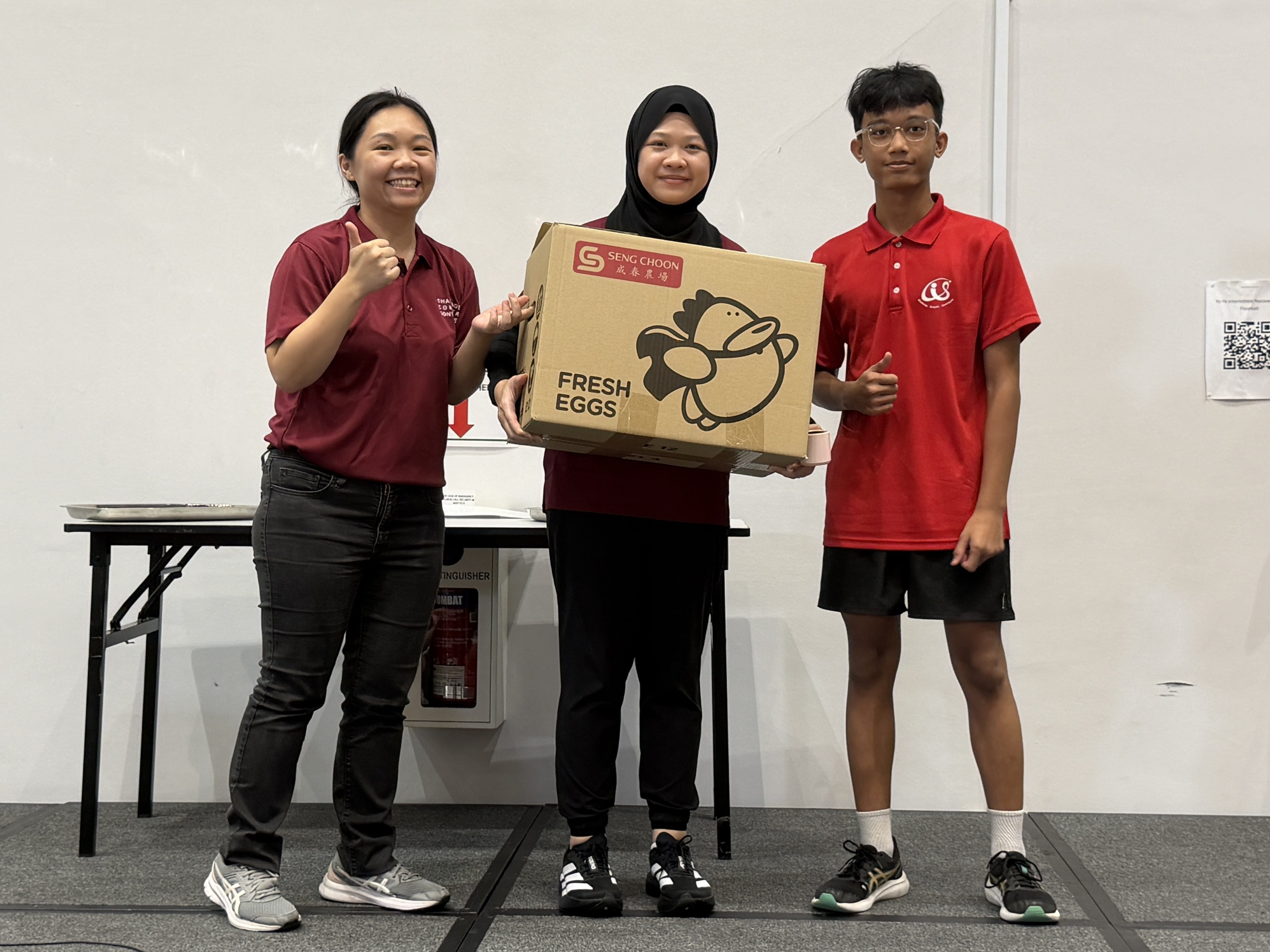 A teacher in a maroon polo shirt and a woman in a hijab flank a young male student in a red school polo shirt, all three holding a Seng Choon Fresh Eggs cardboard box.