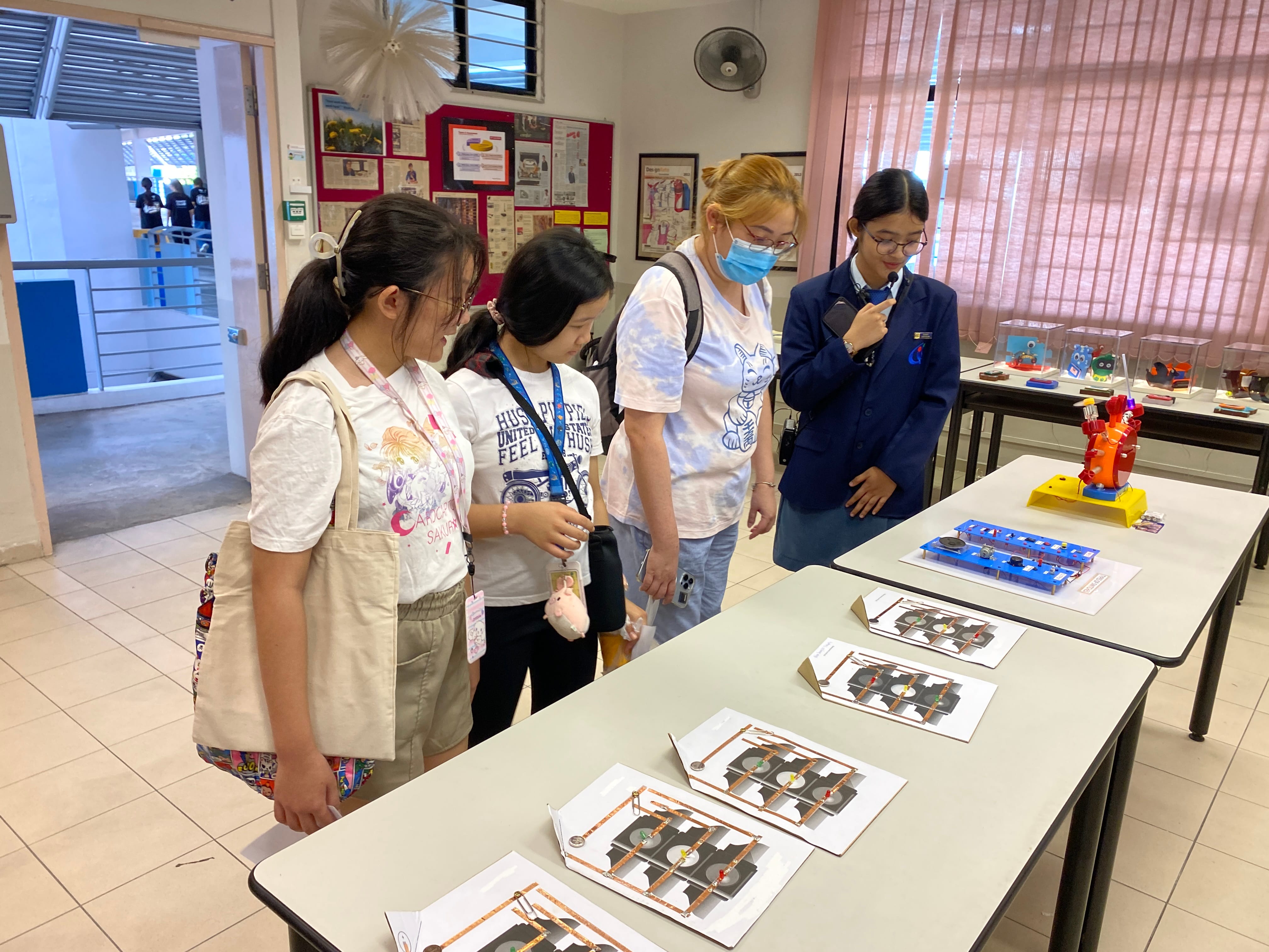 Four students stand at a table examining circuit board displays and colorful models in a classroom exhibit.