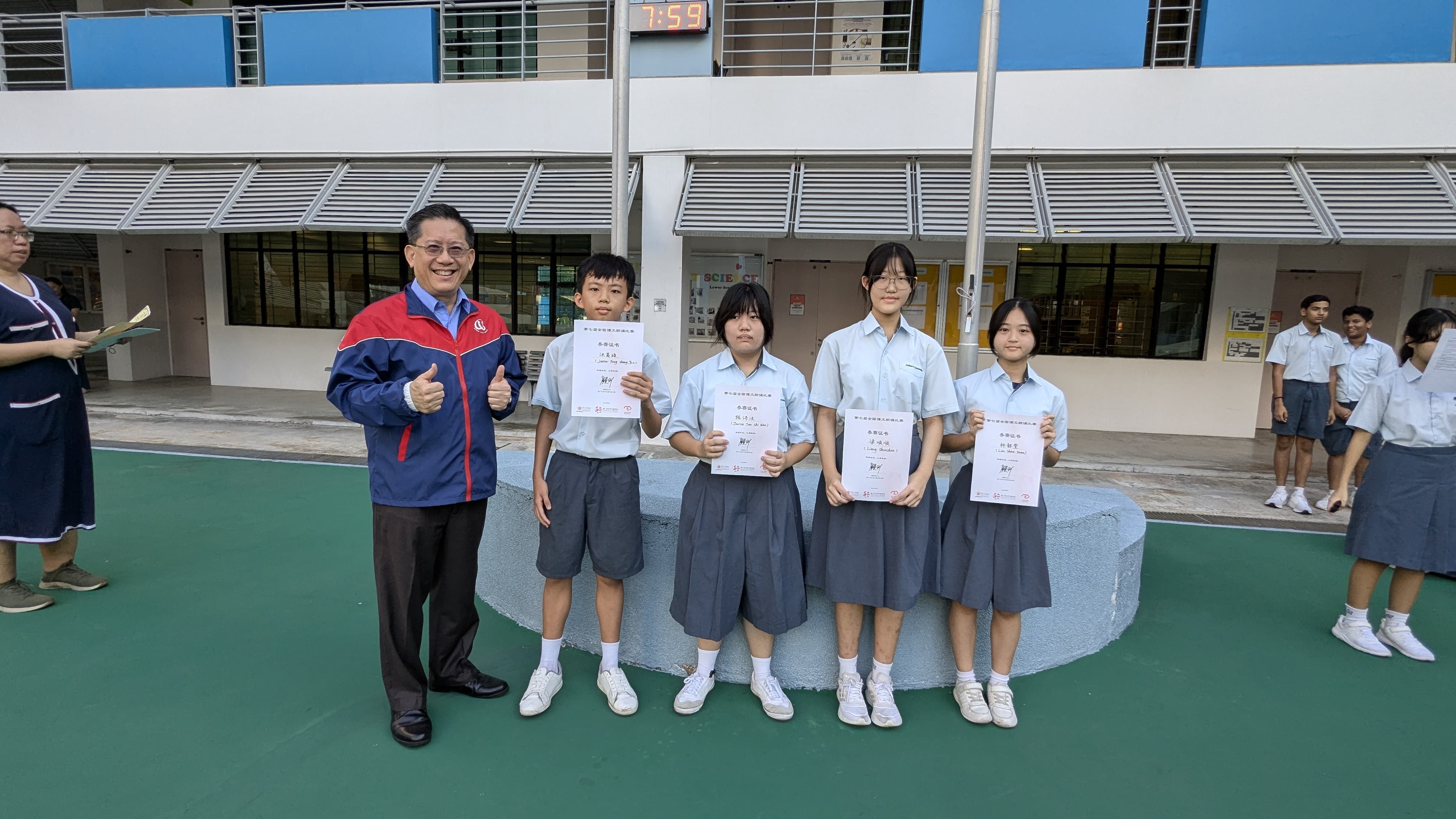 An adult in a red jacket gives thumbs up beside four students in school uniforms holding certificates outdoors.