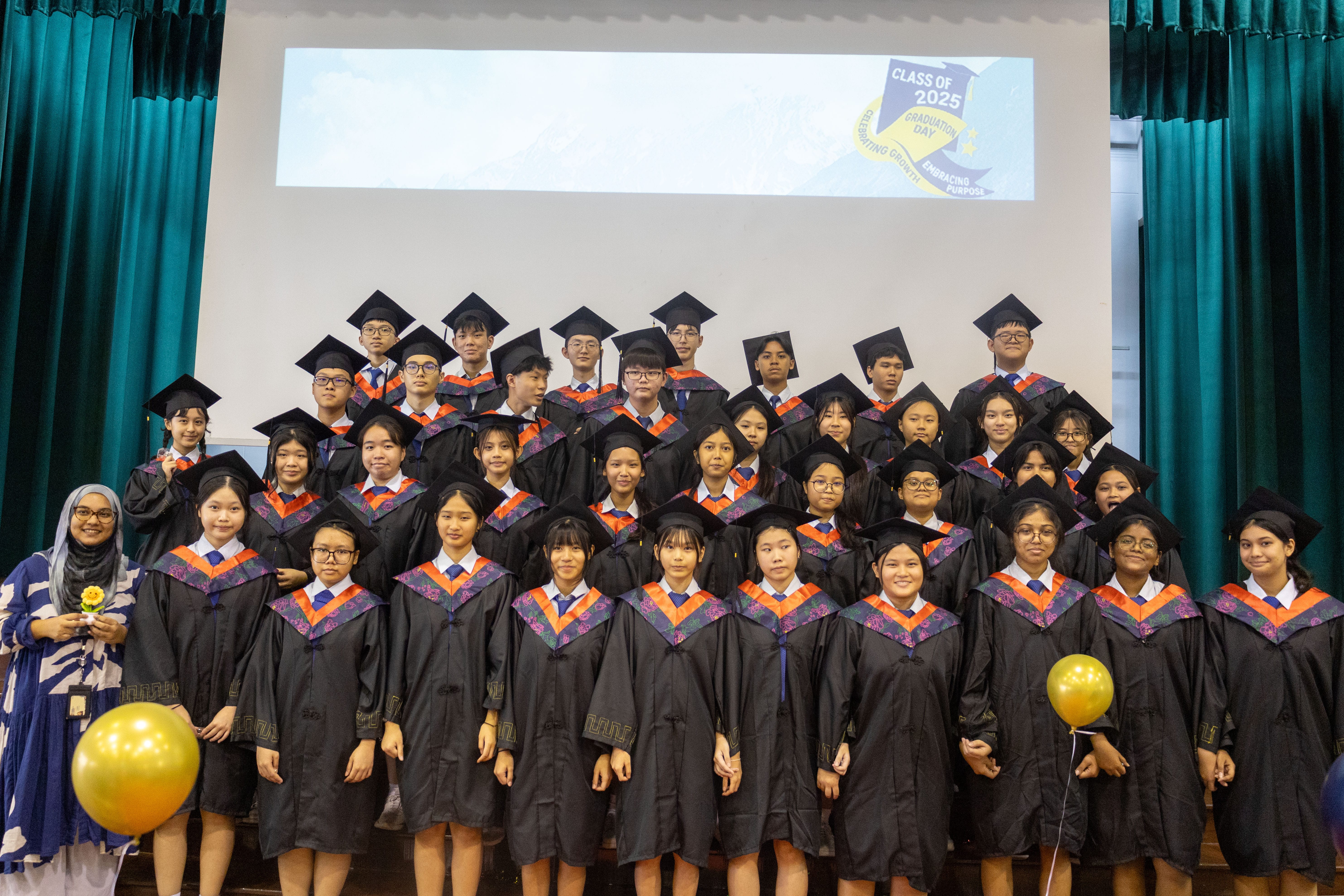 Group of graduates in black caps and gowns on stage under a Class of 2025 banner, with balloons.