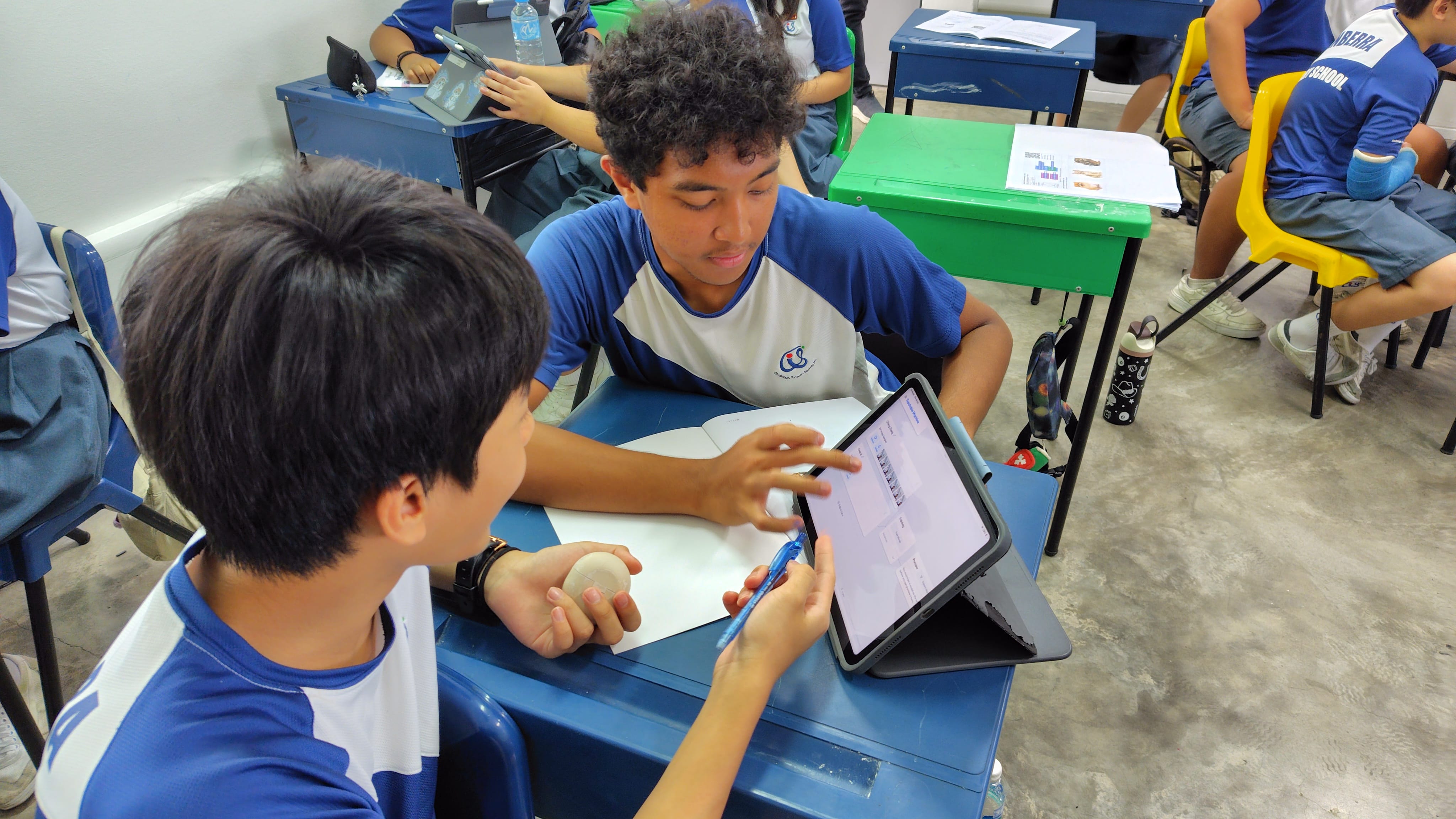 Two students in blue uniforms collaborate over a tablet at a blue desk in a classroom.