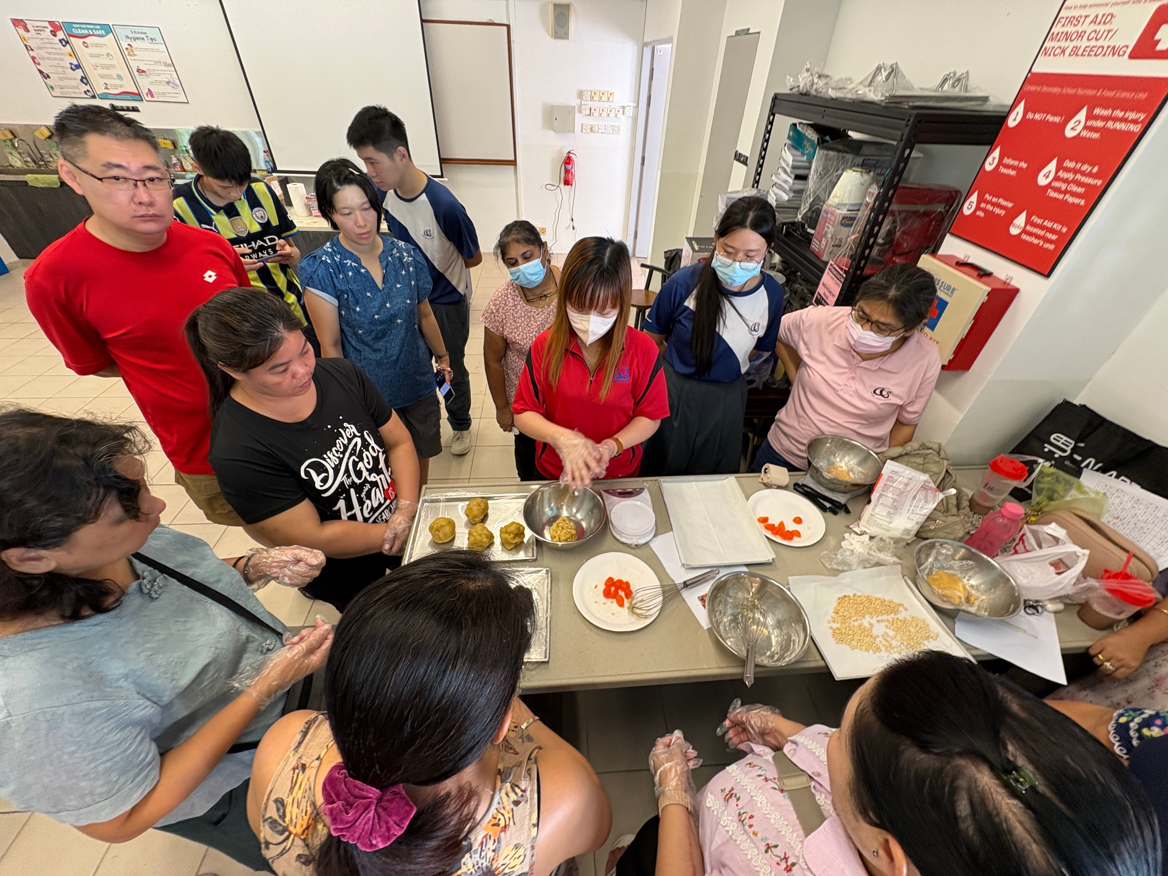 A group of people in a classroom gather around a table during a hands-on cooking session, some wearing masks and gloves.