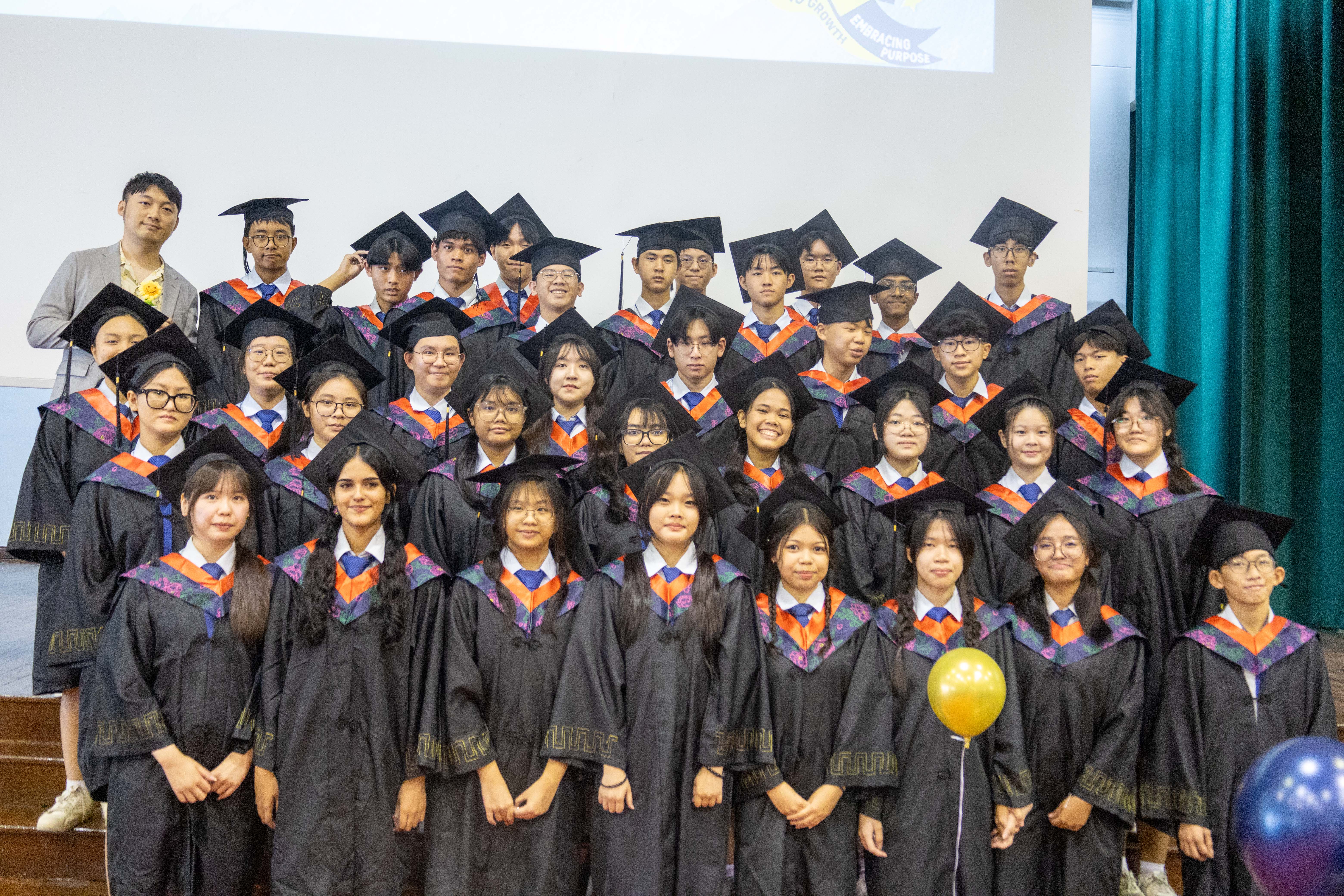Large group of students in black graduation gowns and caps with orange trim posing together indoors.