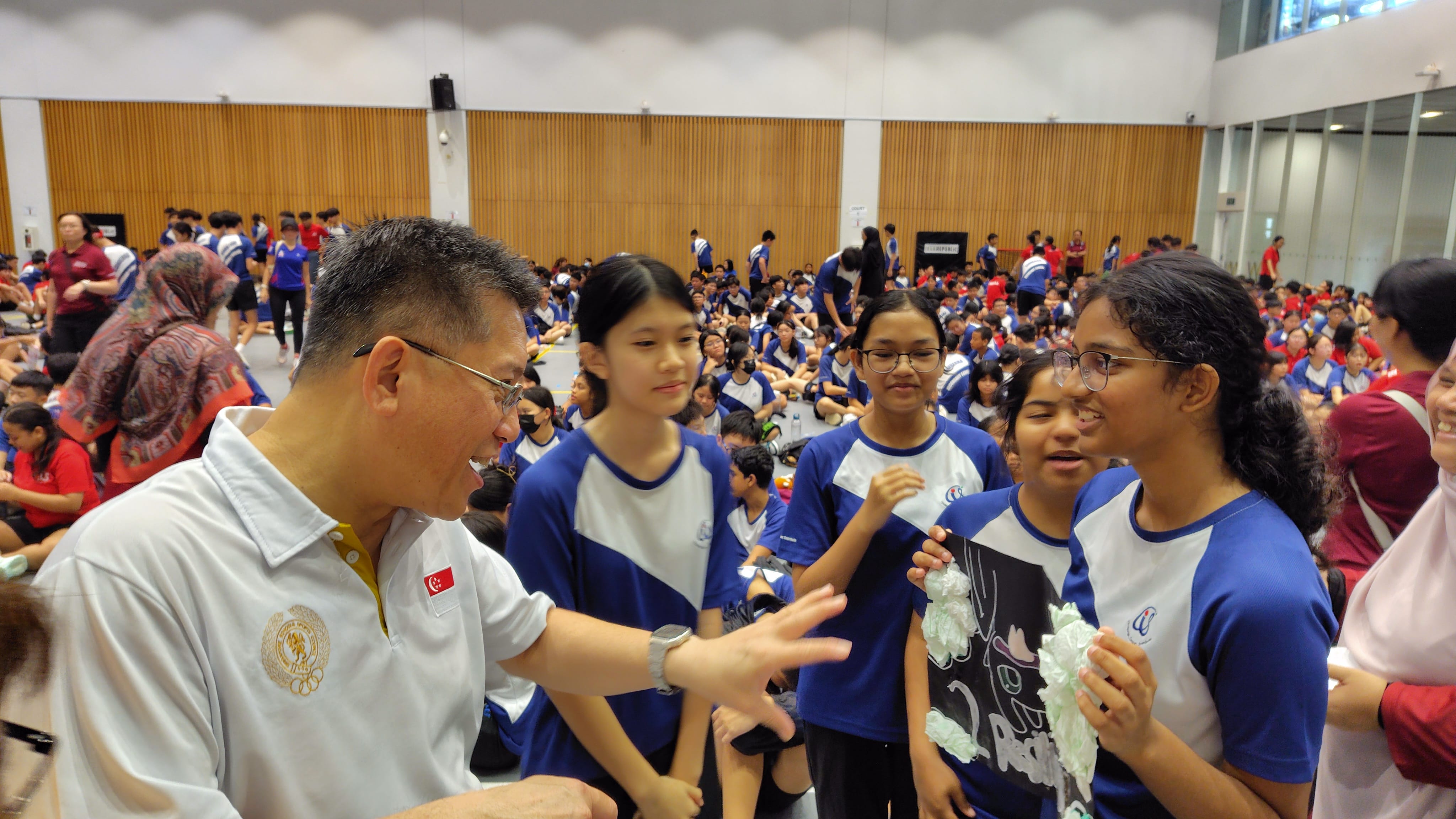 A man in a white polo with a Singapore flag badge talks with several students in blue uniforms inside a crowded hall.