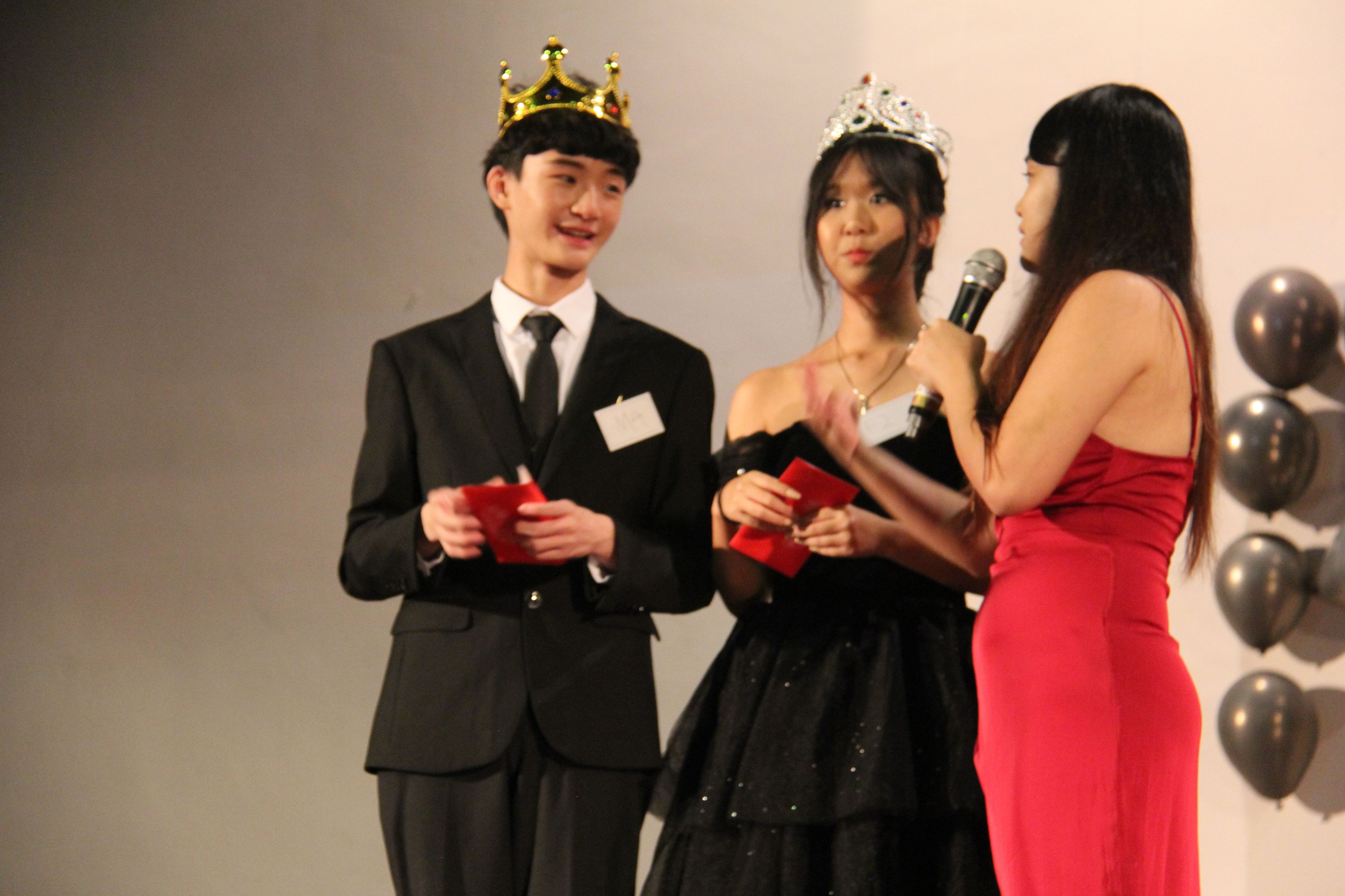 A young man in a suit with a gold crown and a woman in a black gown with a silver tiara stand on stage.