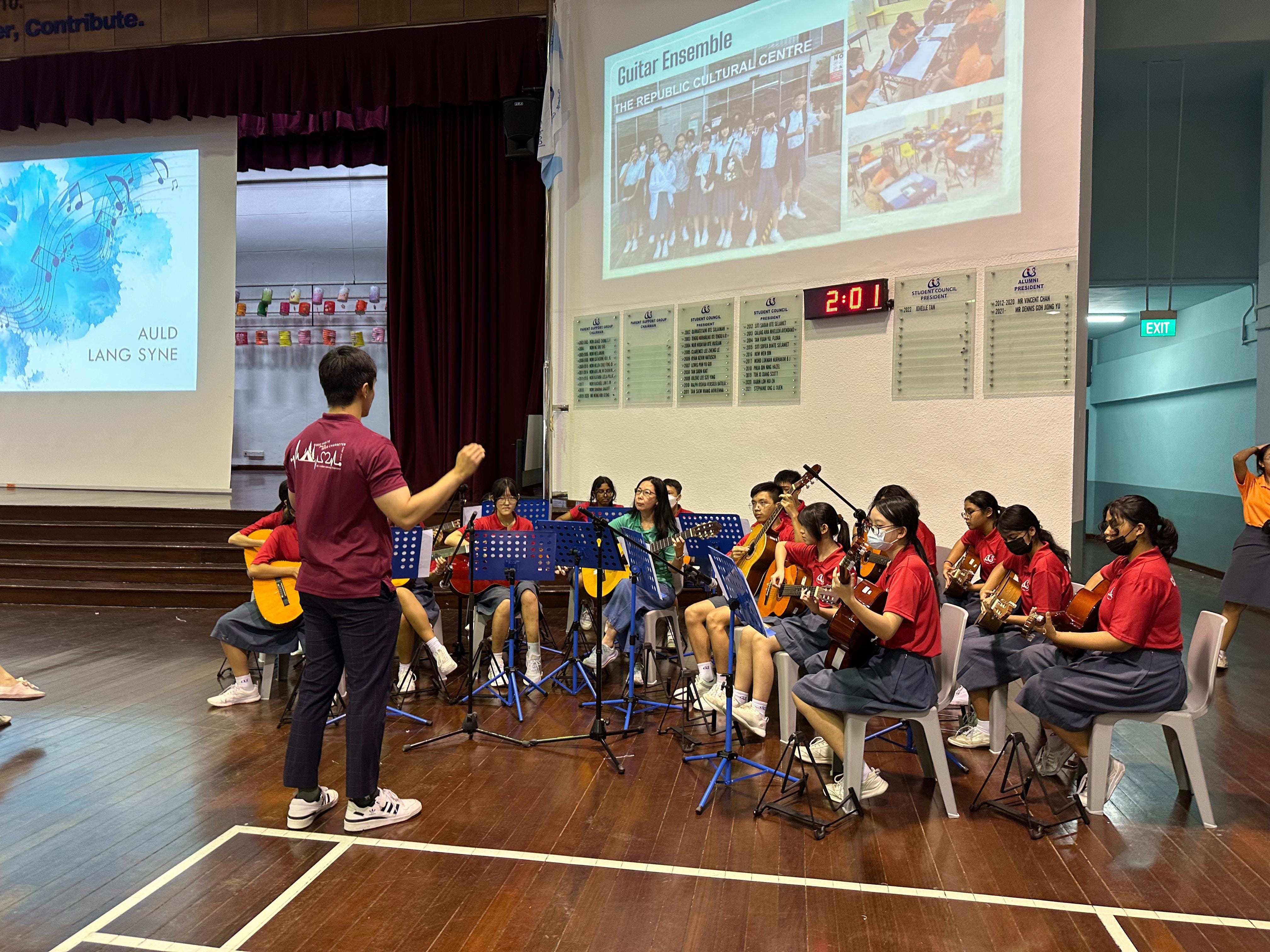 A conductor leads a student guitar ensemble in a school hall with a projected Guitar Ensemble slideshow behind them.