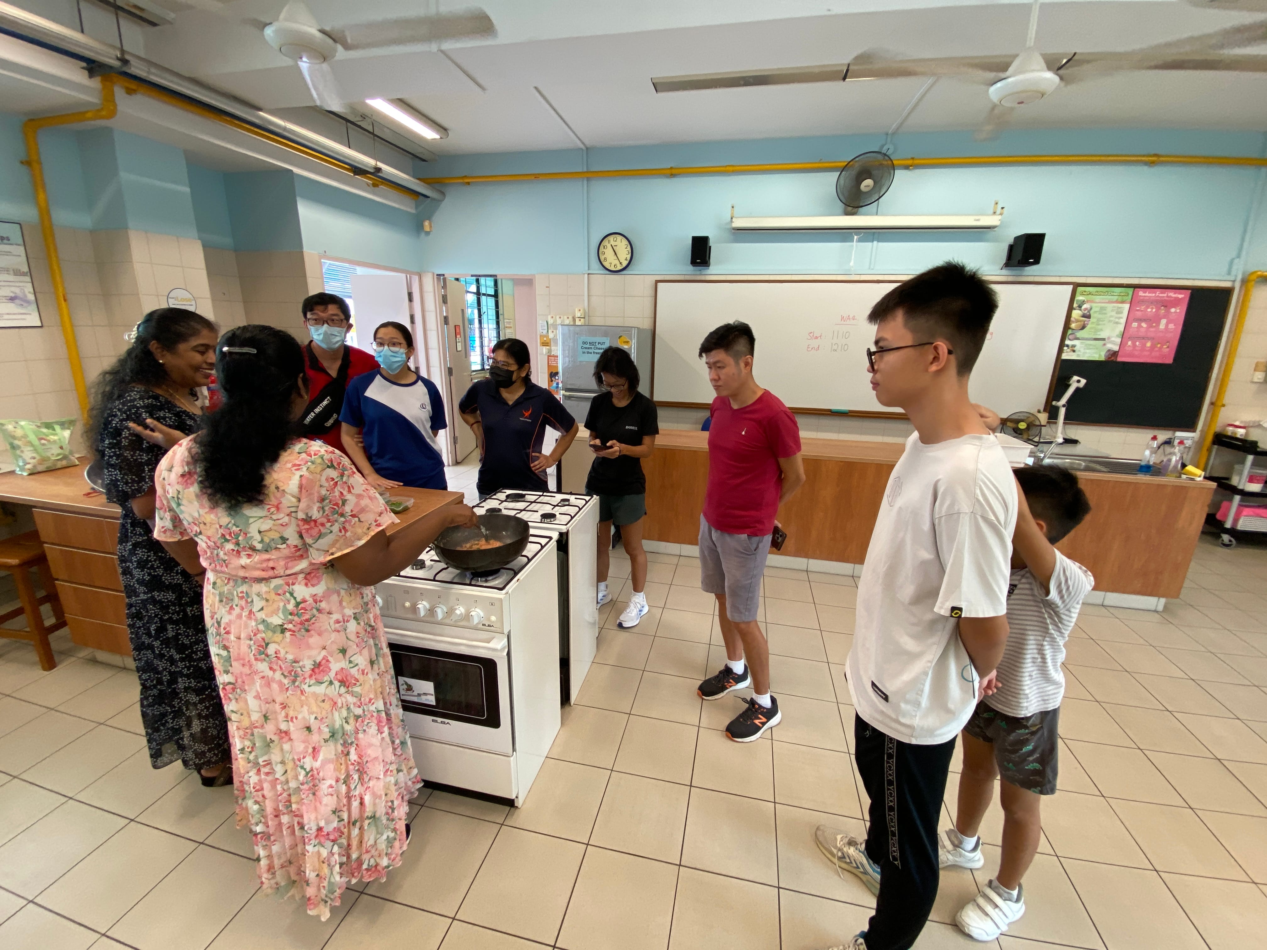 A woman in a floral dress cooks on a gas stove while a group of people watch in a school cooking classroom.