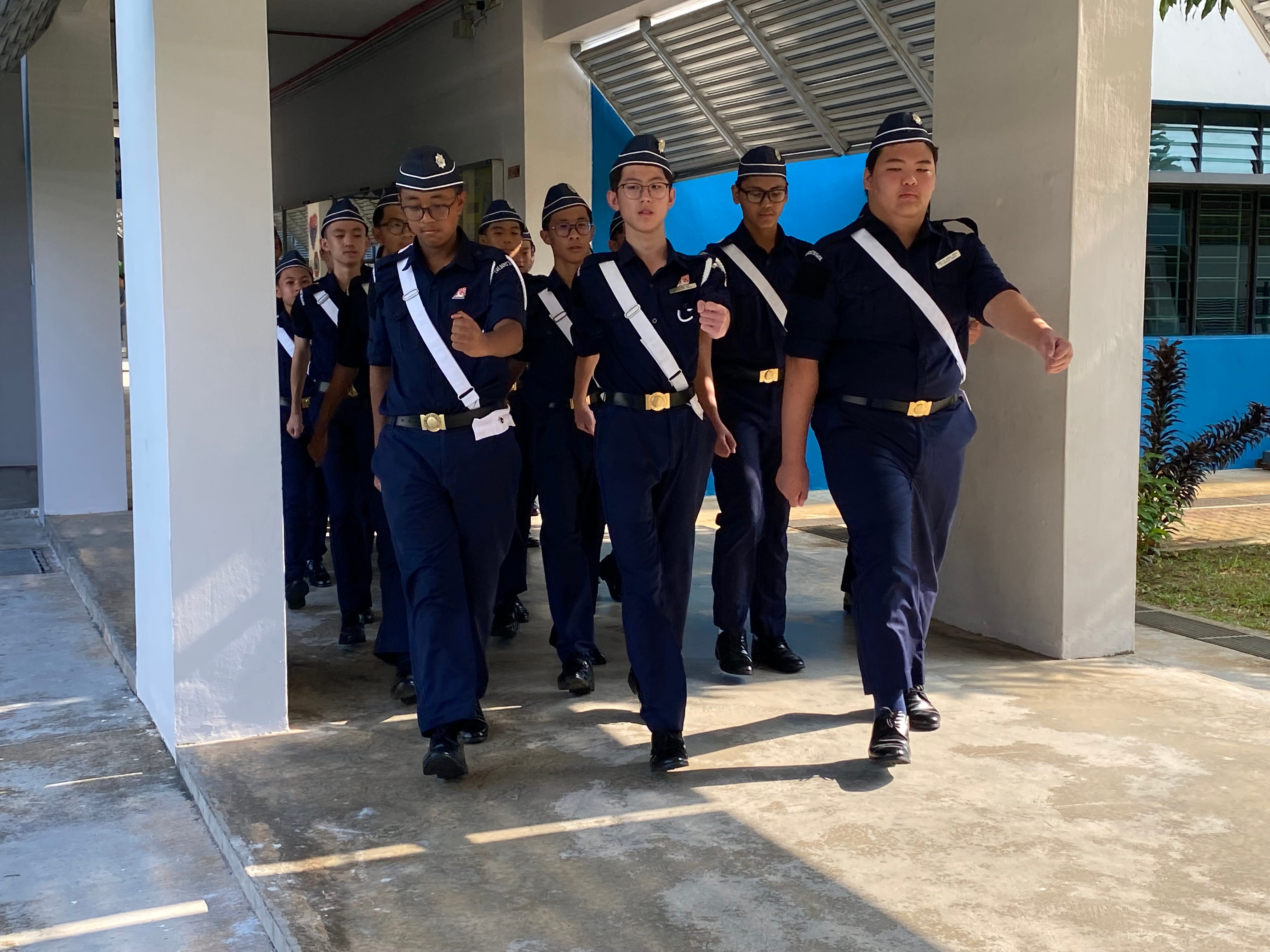 A group of young cadets in navy blue uniforms with white sashes marching in formation under a covered walkway.