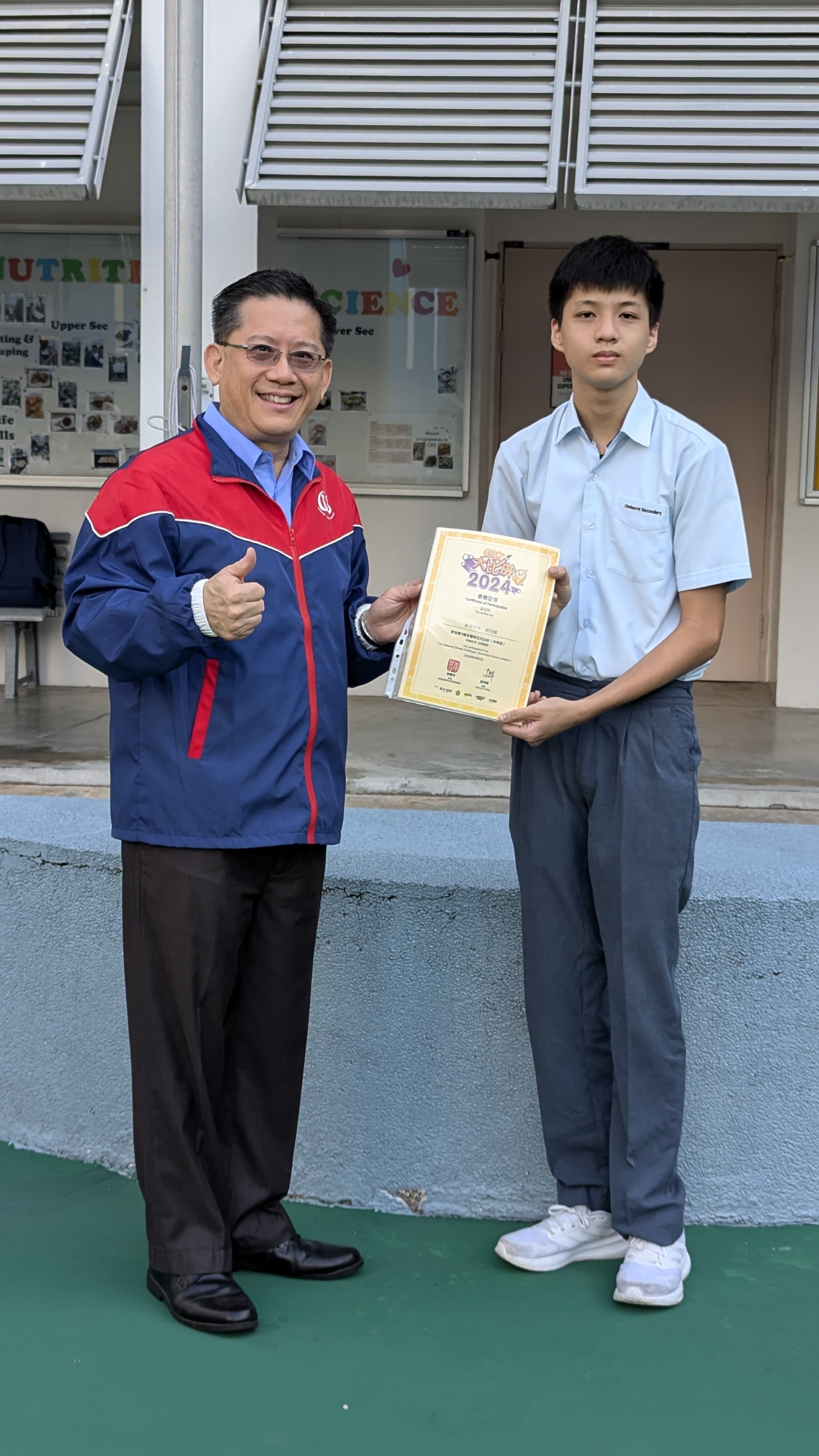 An older man in a red and blue jacket gives a thumbs up beside a student holding a 2024 certificate outdoors.