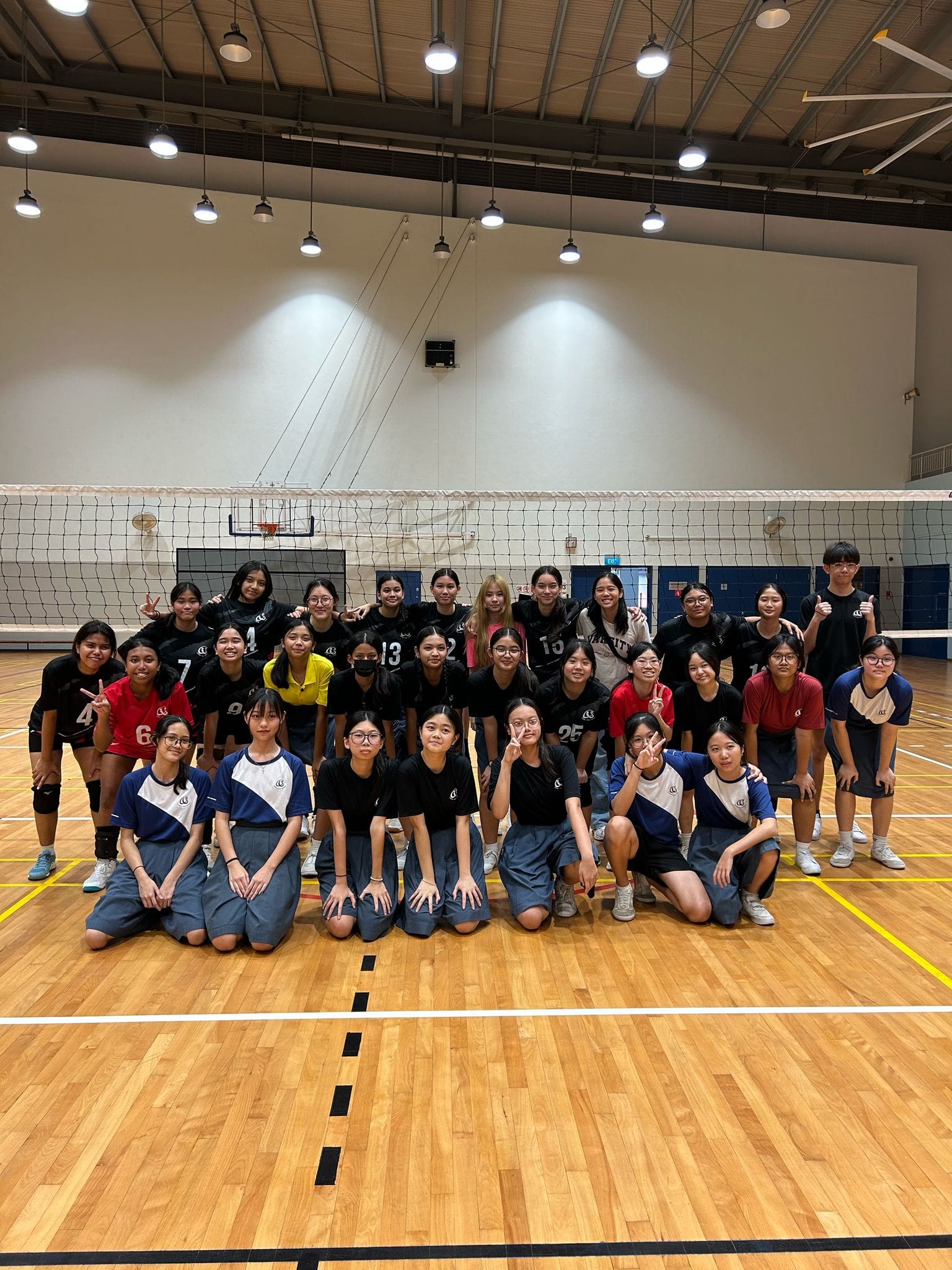 A large group of young volleyball players posing together in front of a net inside an indoor gymnasium.