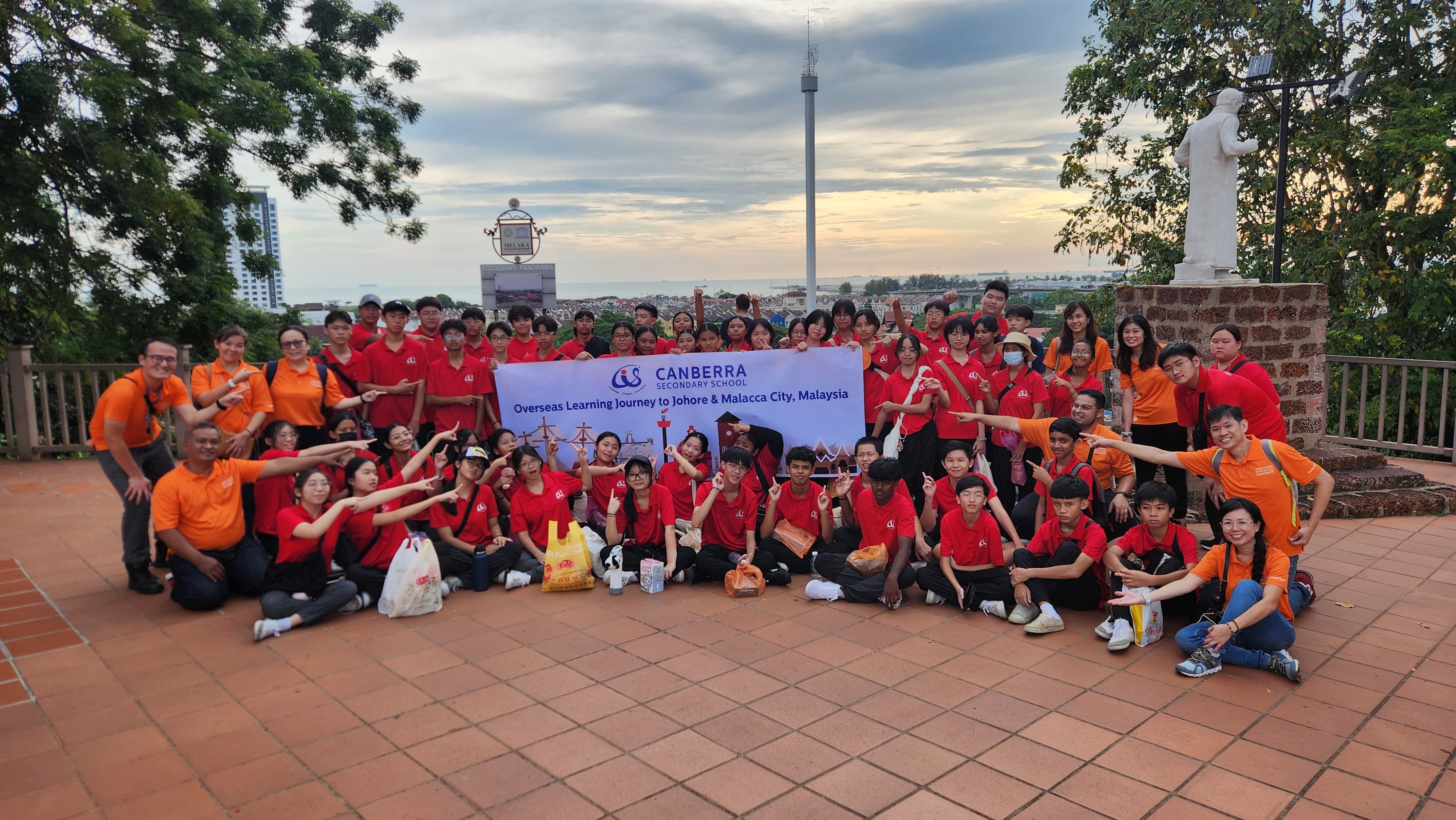Large group of students in red shirts with teachers in orange, holding a Canberra Secondary School Malaysia trip banner.