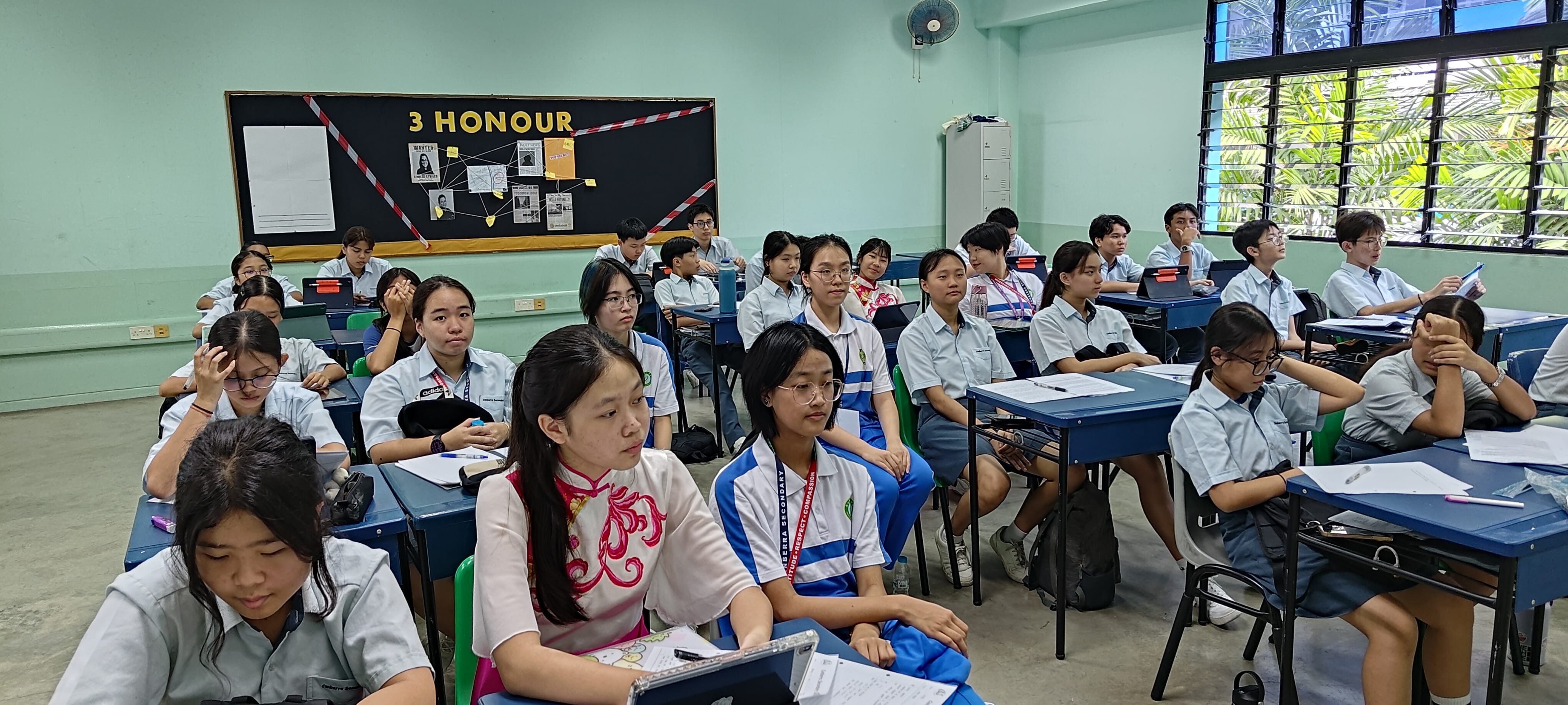 Students in uniforms sit at desks in a classroom labeled "3 HONOUR" on a black bulletin board.