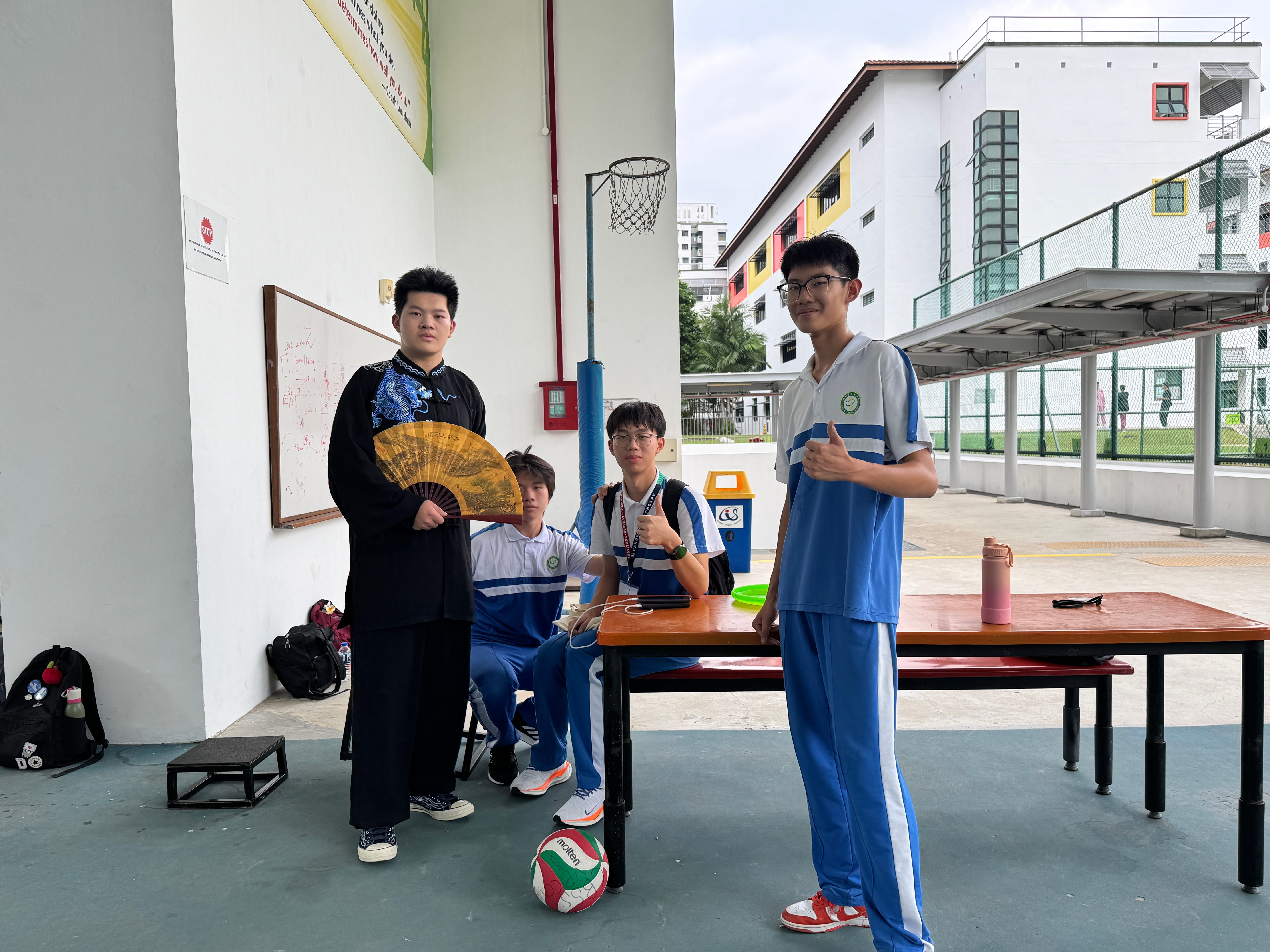 Four students by a table; one in black robe holding a fan, another standing gives thumbs-up, volleyball on floor.