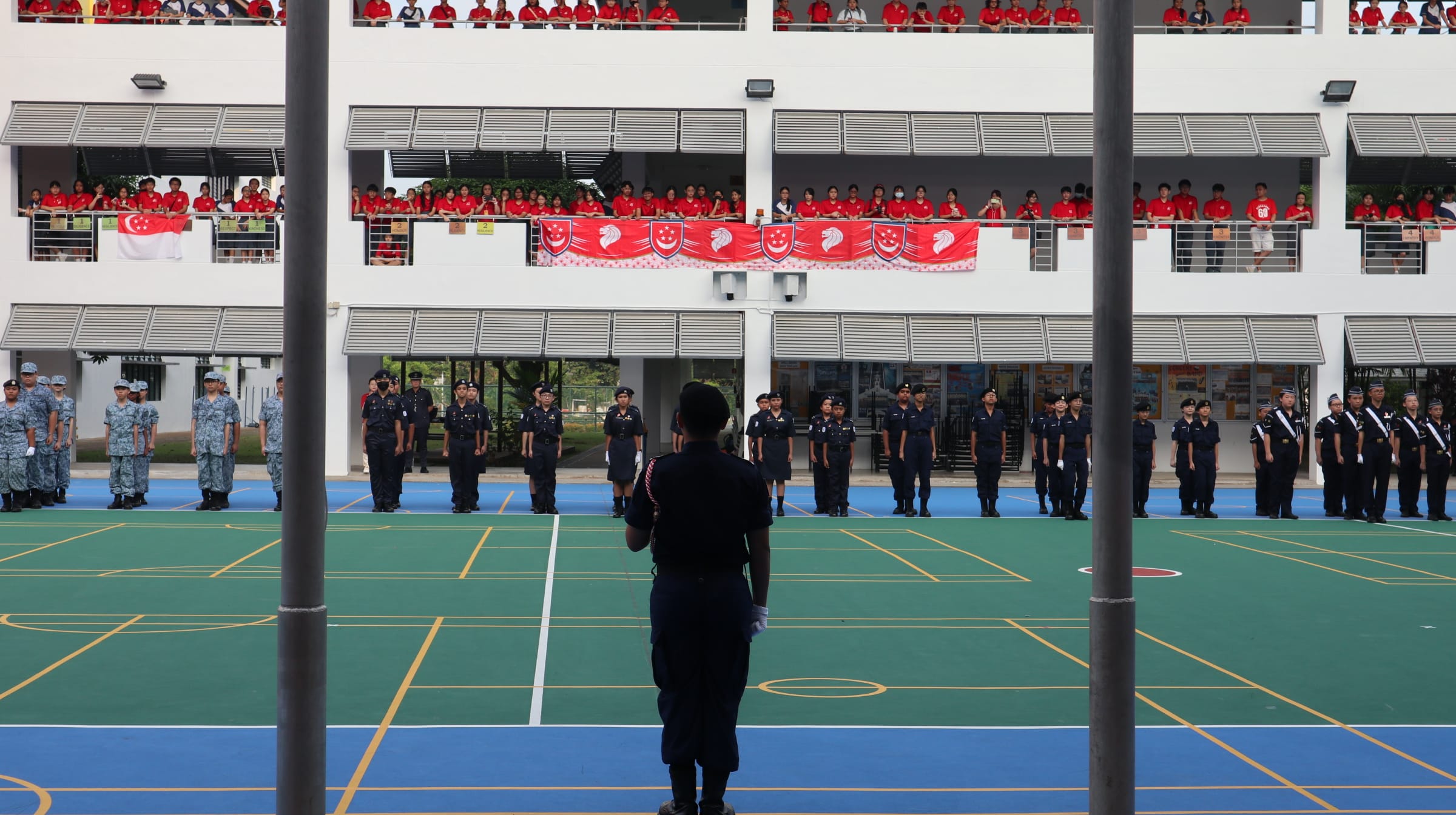 uniformed cadets standing in formation on a school court while students in red watch from building balconies above